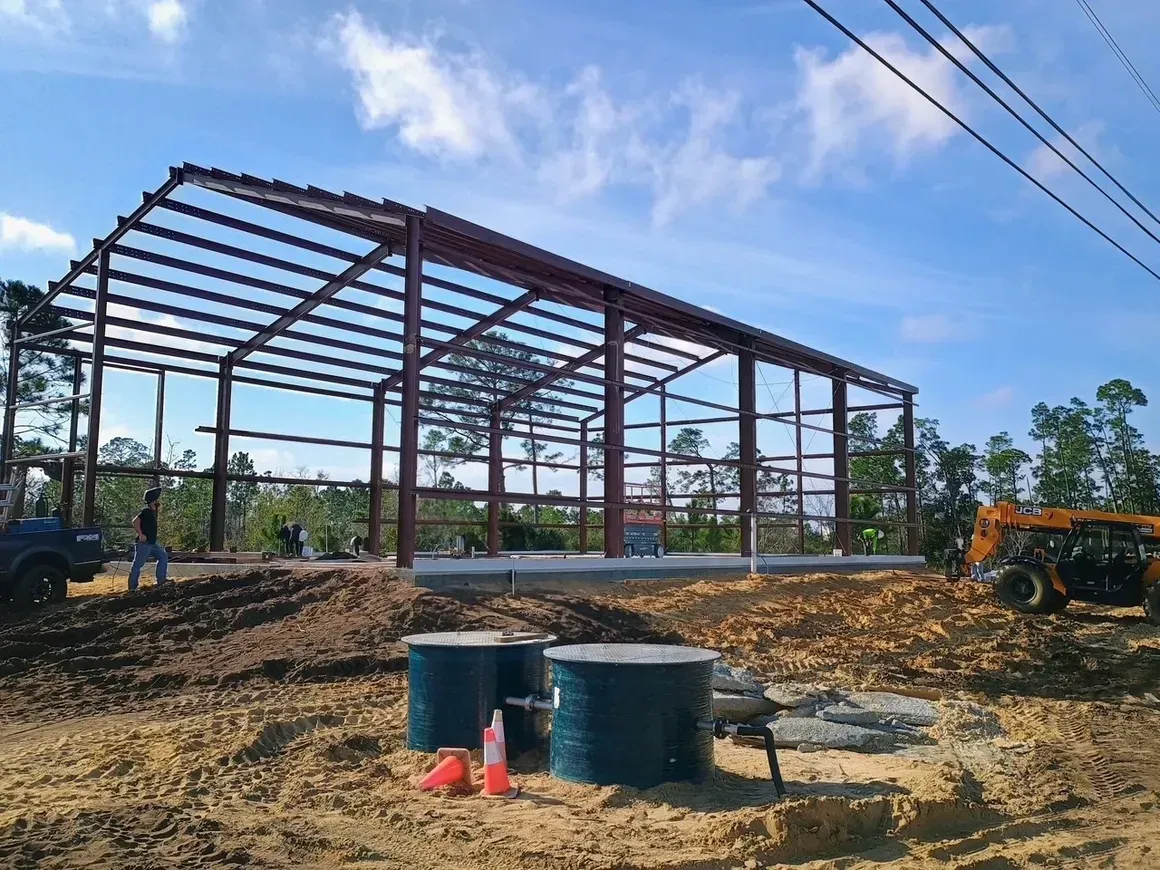 Steel frame building under construction on a muddy site, with barrels, equipment, and excavators nearby