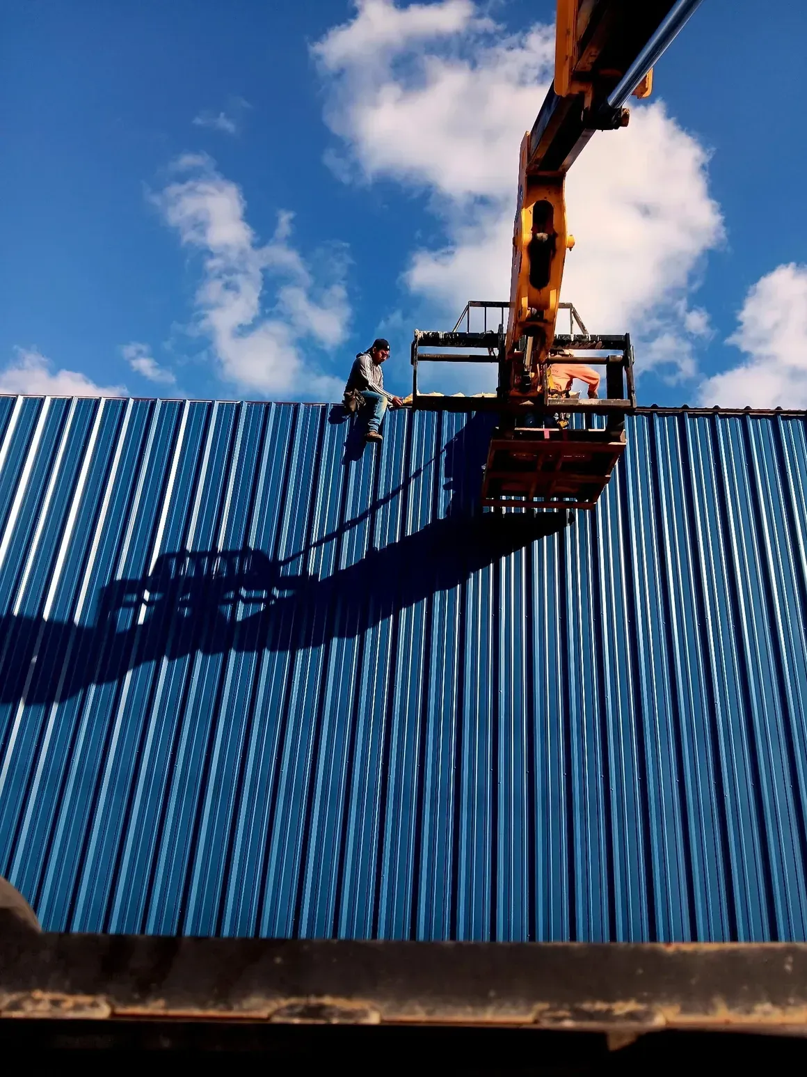 Worker on a blue roof using a crane lift against a cloudy sky