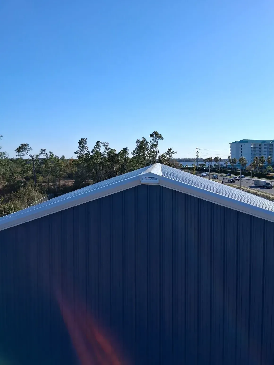 Blue metal roof peak against a clear sky, with trees and a building in the background.