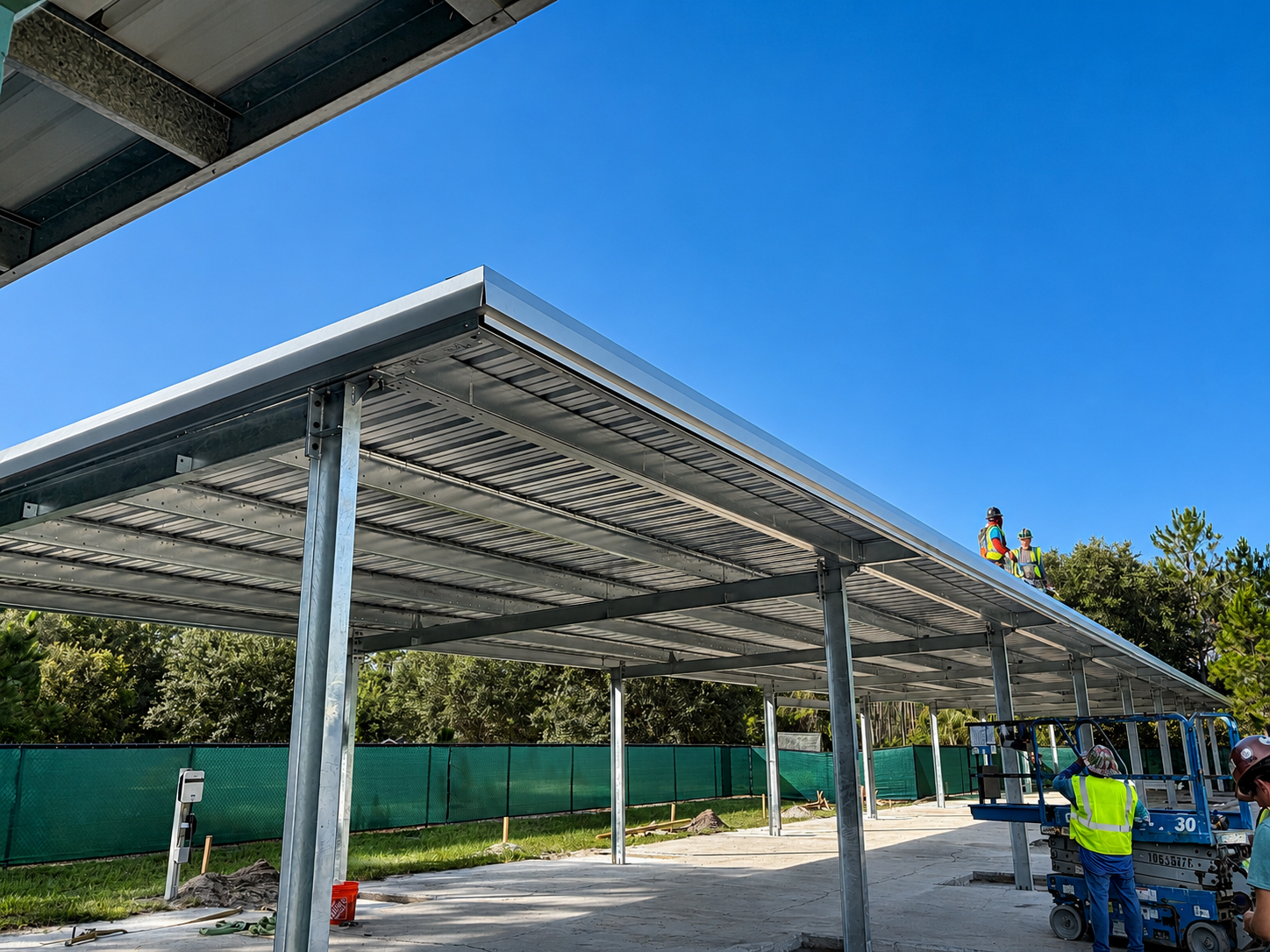 Workers install a metal carport frame under a bright blue sky.