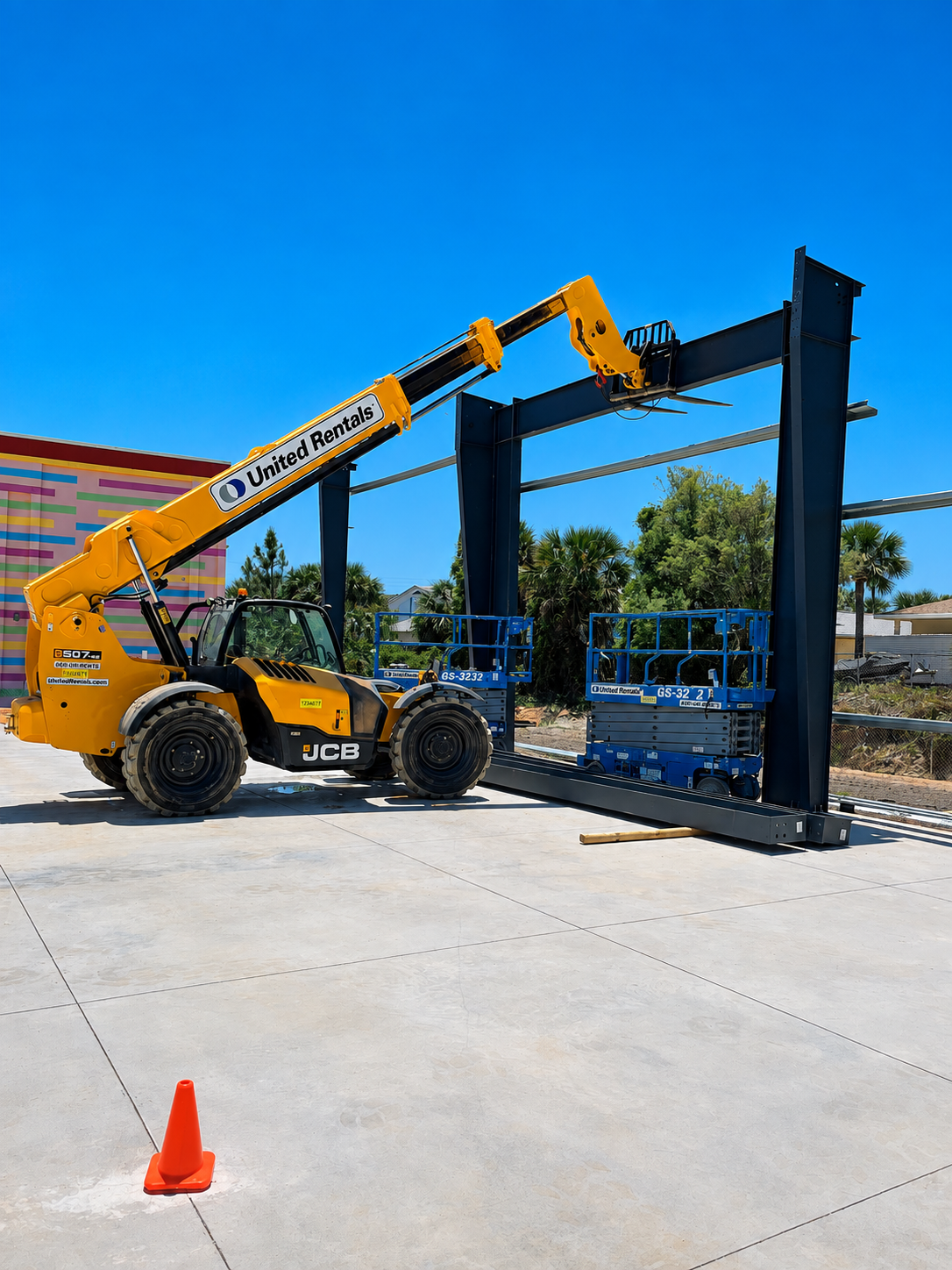 Yellow telescopic forklift lifting a blue metal frame in a sunny outdoor yard with an orange cone.