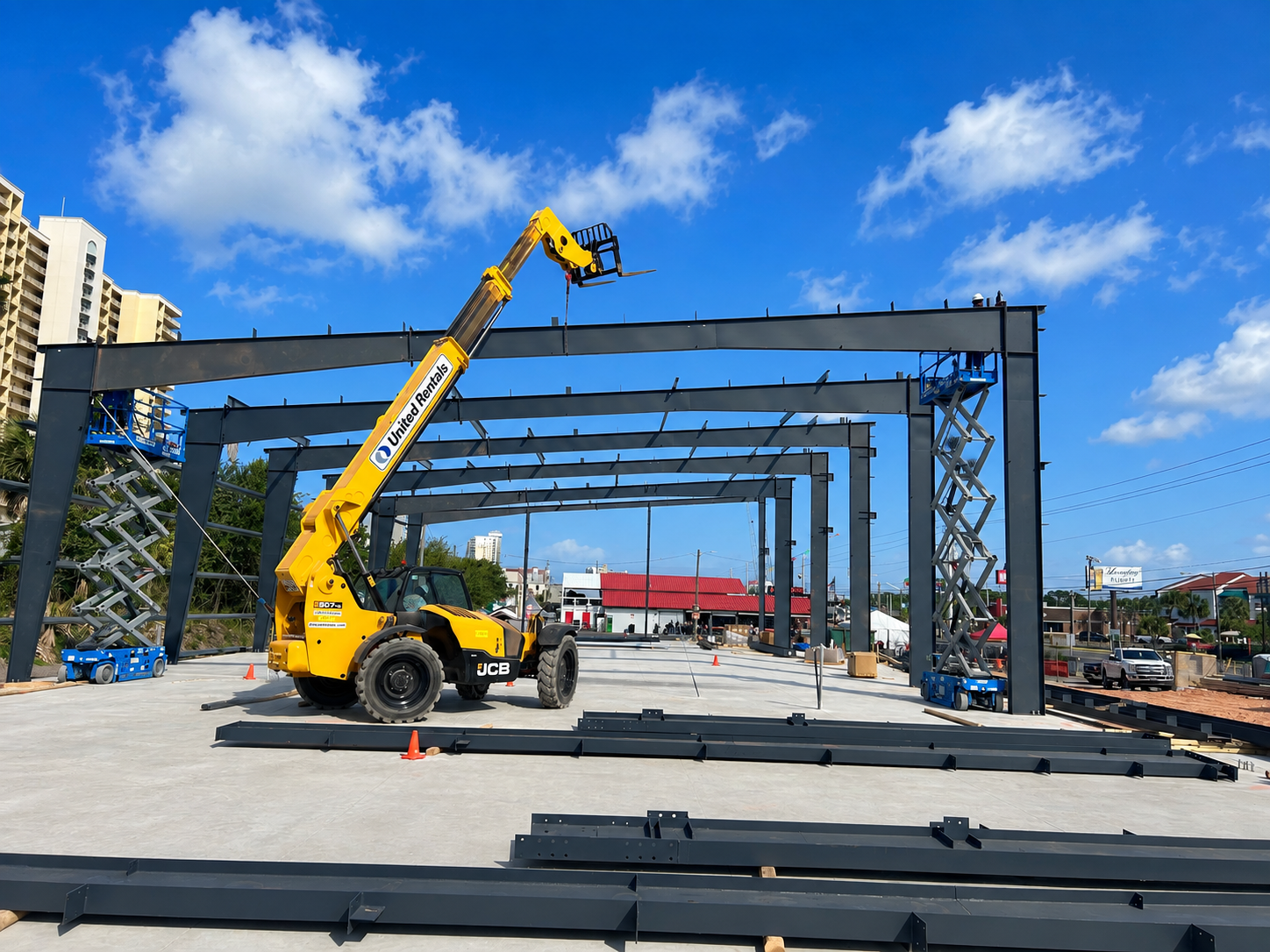 Construction site with yellow crane lifting steel beams for a large frame structure under a blue sky.