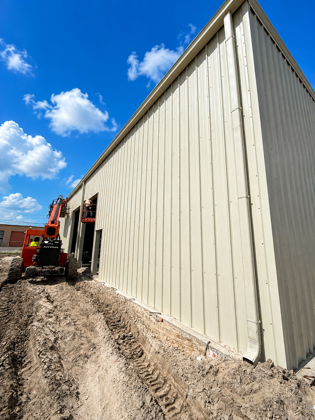 Long white metal building under construction beside a muddy dirt path and blue sky, with a red lift at the left.