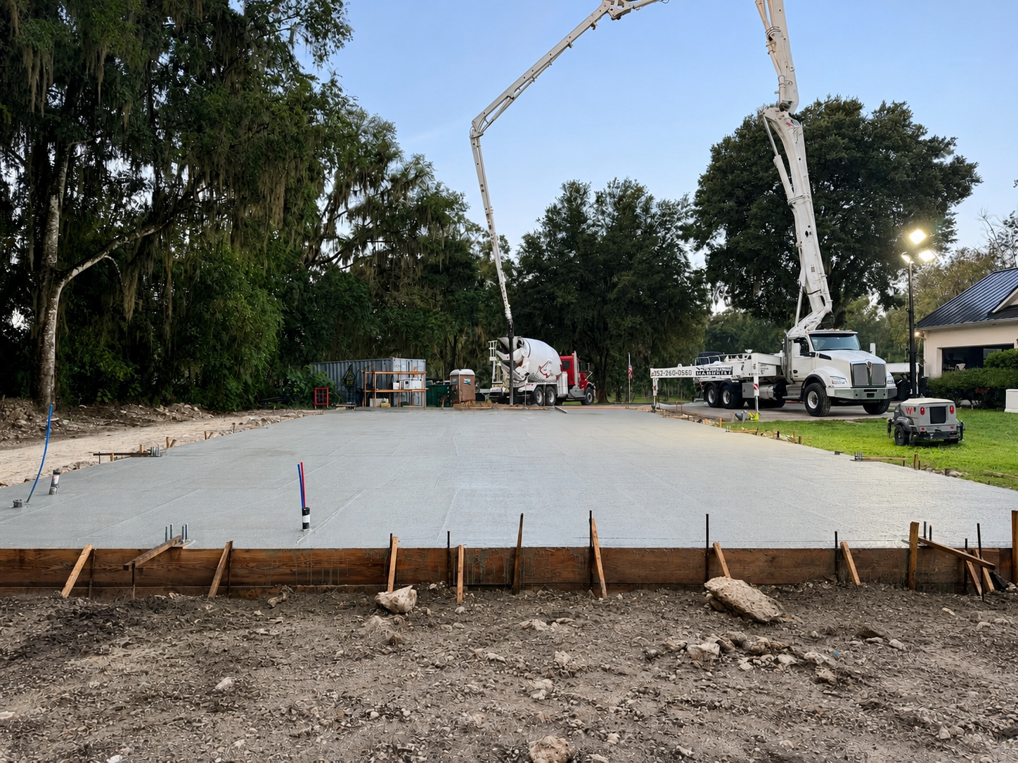 Concrete slab being poured at a construction site, with workers, a pump truck, and trees in the background