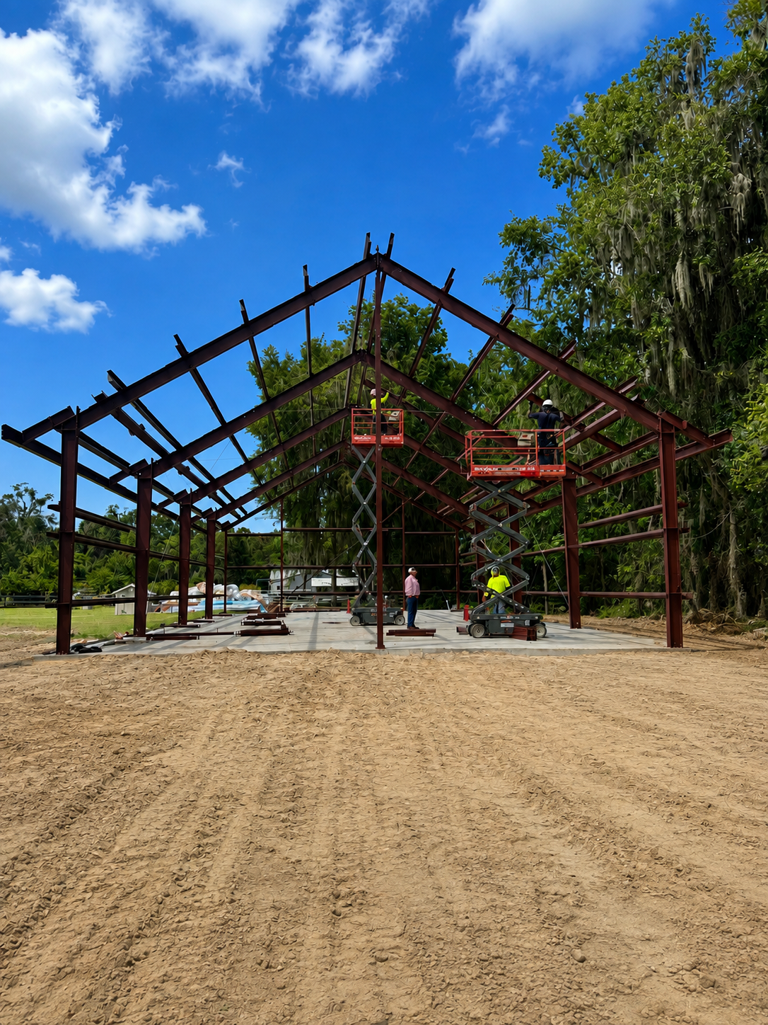Open-air pavilion under construction with red steel frame, dirt foreground, and blue sky with clouds