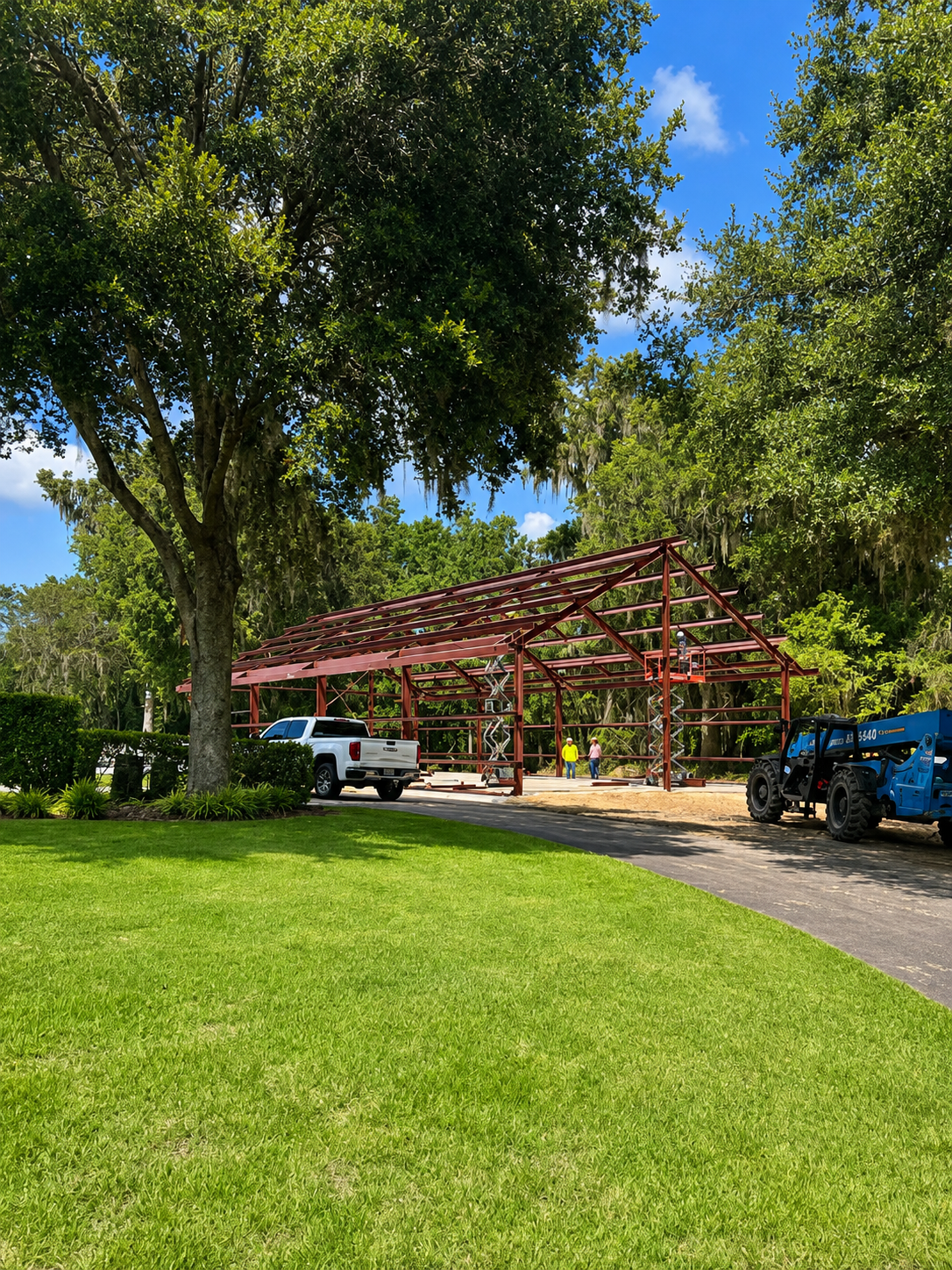 Park with green lawn, large trees, and a red steel pavilion beside parked vehicles on a sunny day