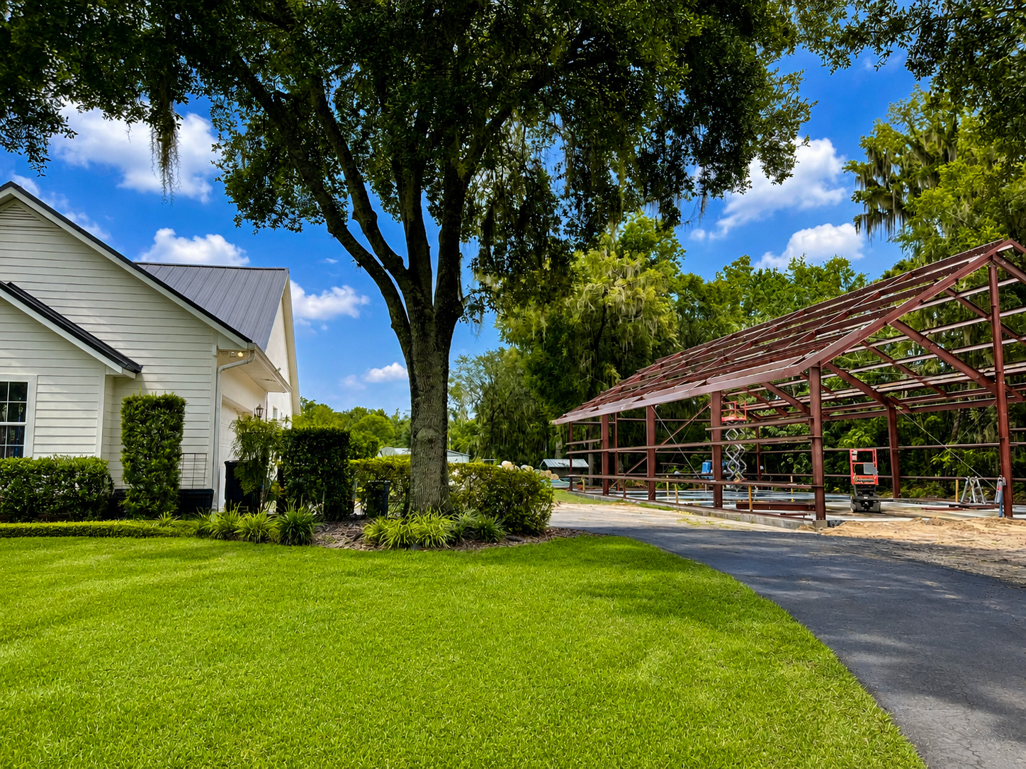 Suburban driveway beside a white house, green lawn, large trees, and a red carport under a blue sky