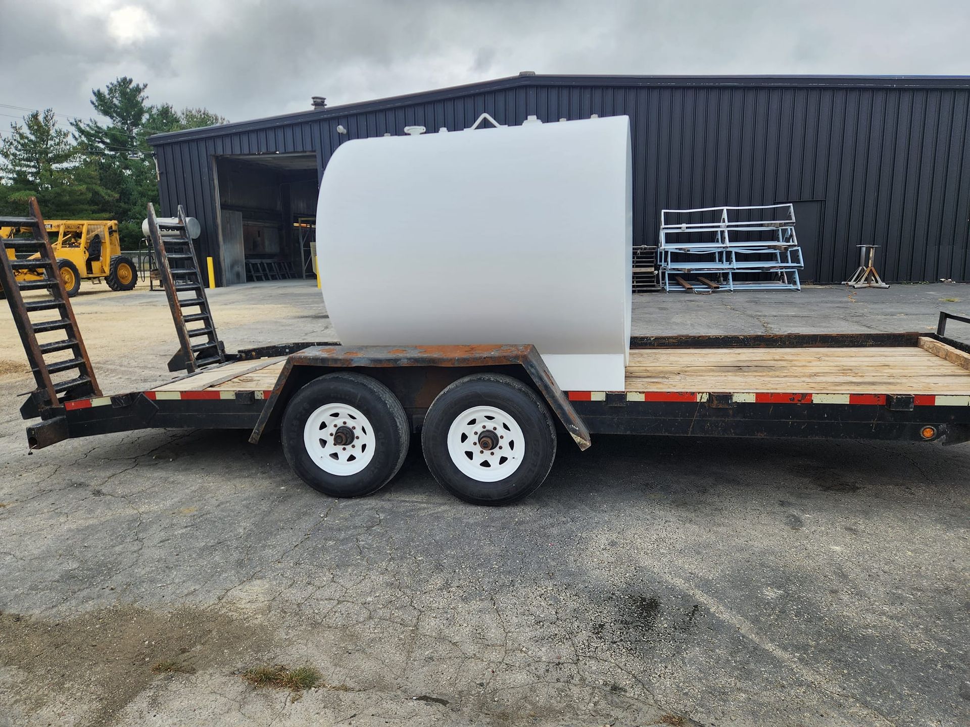 A white cylindrical tank mounted on a flatbed equipment trailer in a gravel lot outside a warehouse.
