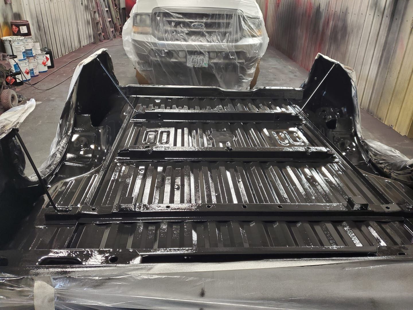 A freshly painted black truck bed in a workshop, with a plastic-covered vehicle visible in the background.