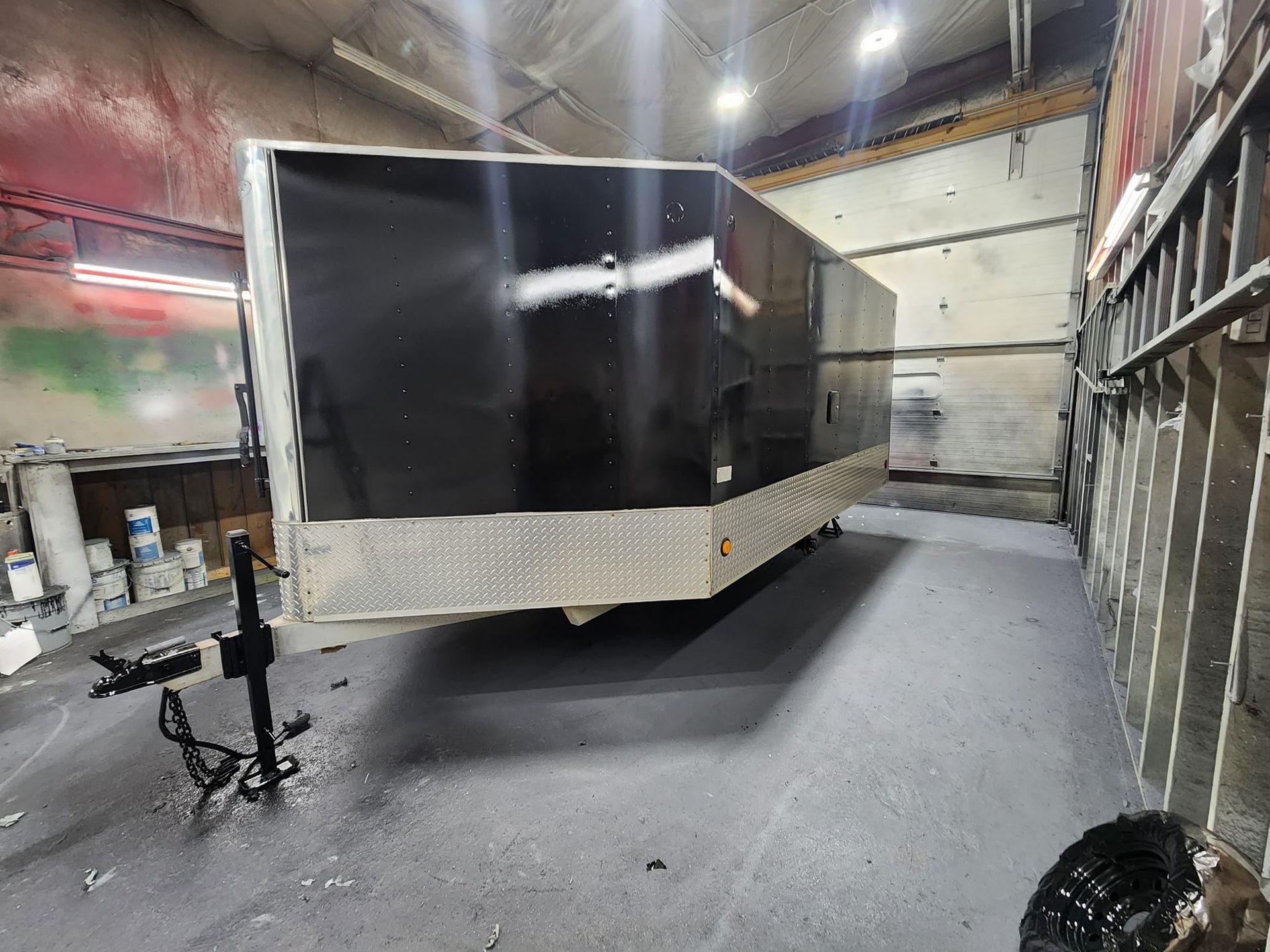 A black enclosed trailer with a diamond-plate lower panel parked inside a workshop with overhead lighting.
