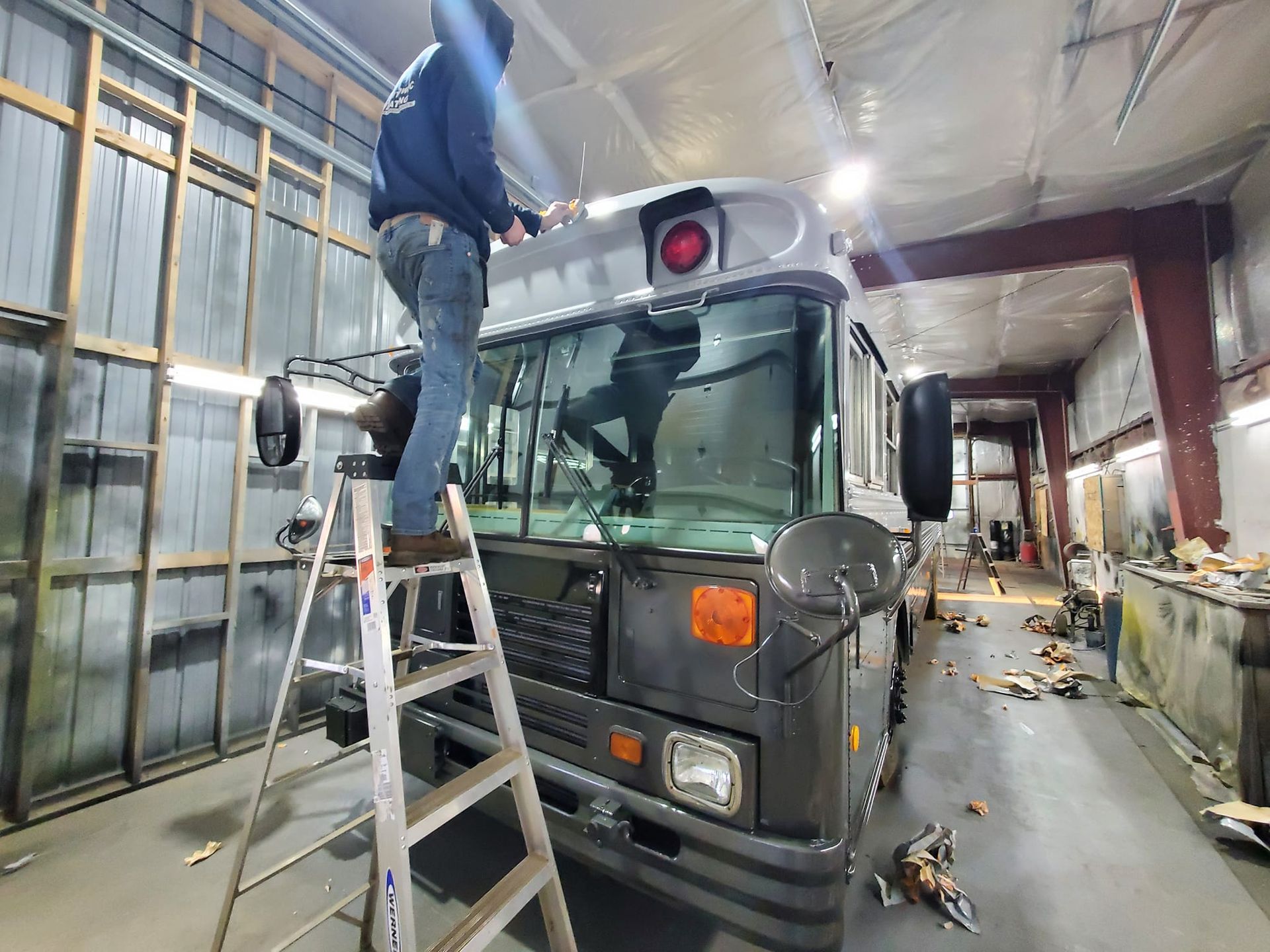 A person stands on a step ladder working on the roof of a grey bus inside a workshop.