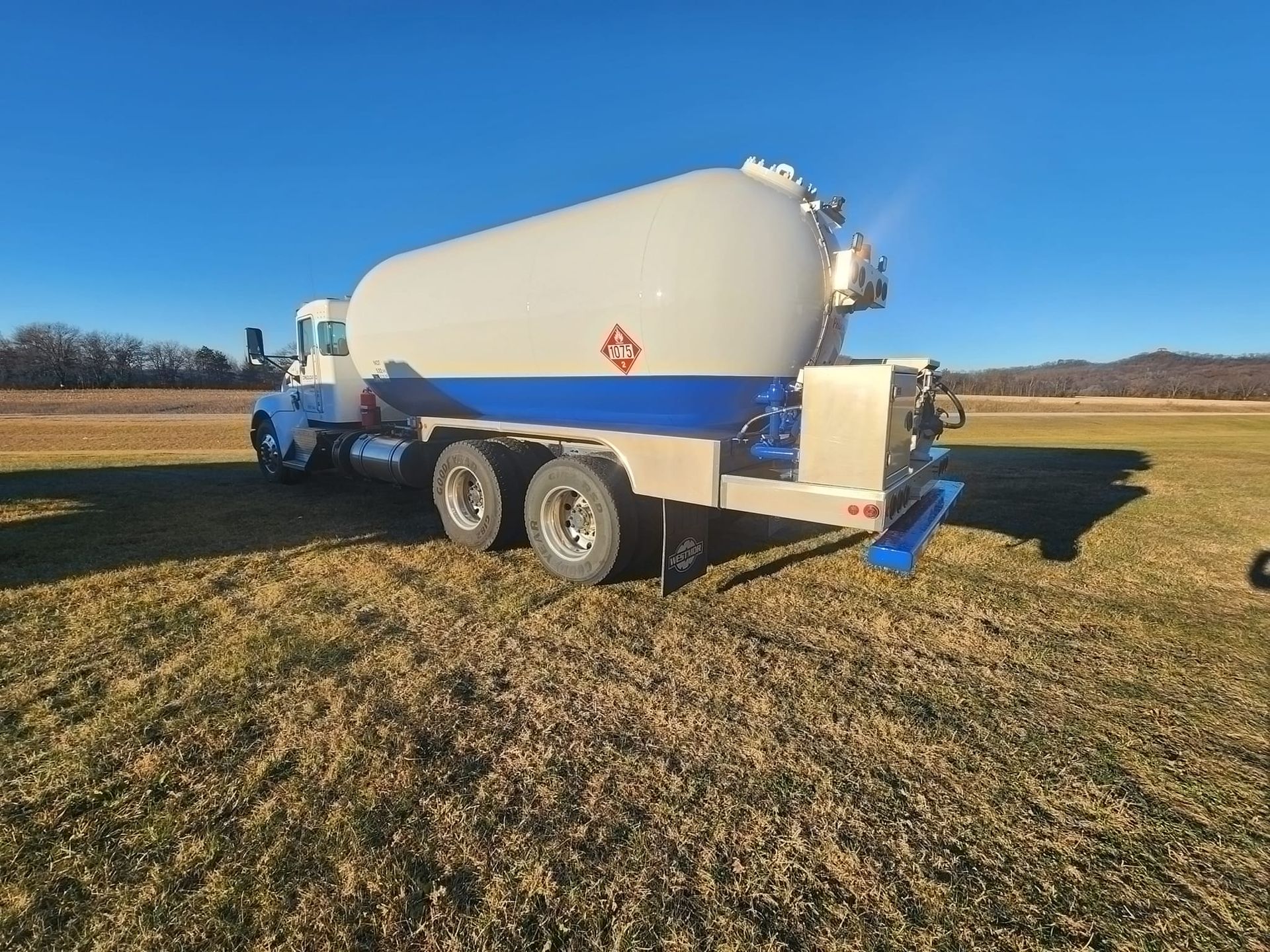 A white and blue propane delivery truck parked in a large, dry grassy field under a clear blue sky.