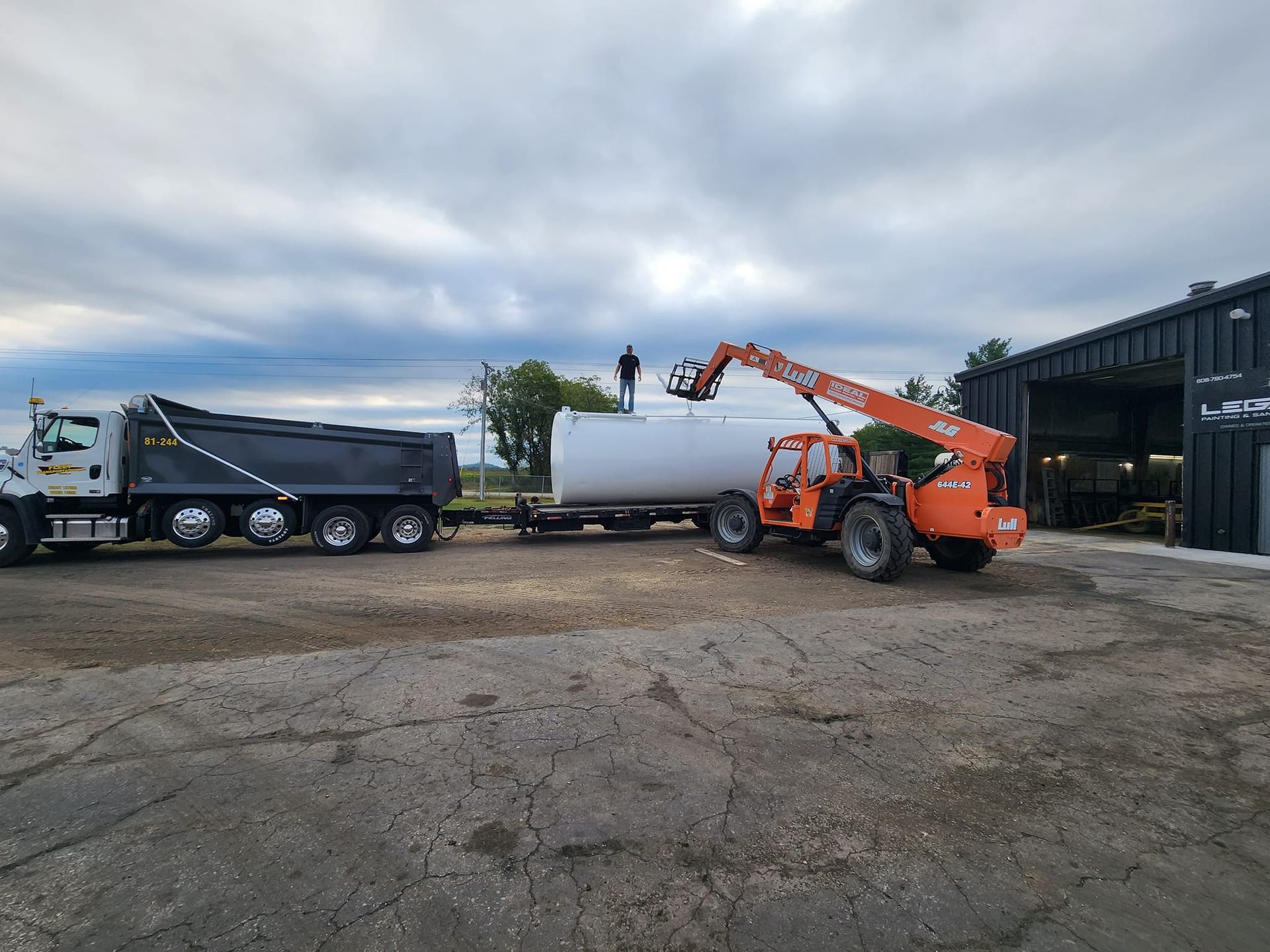 A person stands atop a large white cylindrical tank on a trailer, next to a dump truck and an orange telehandler.