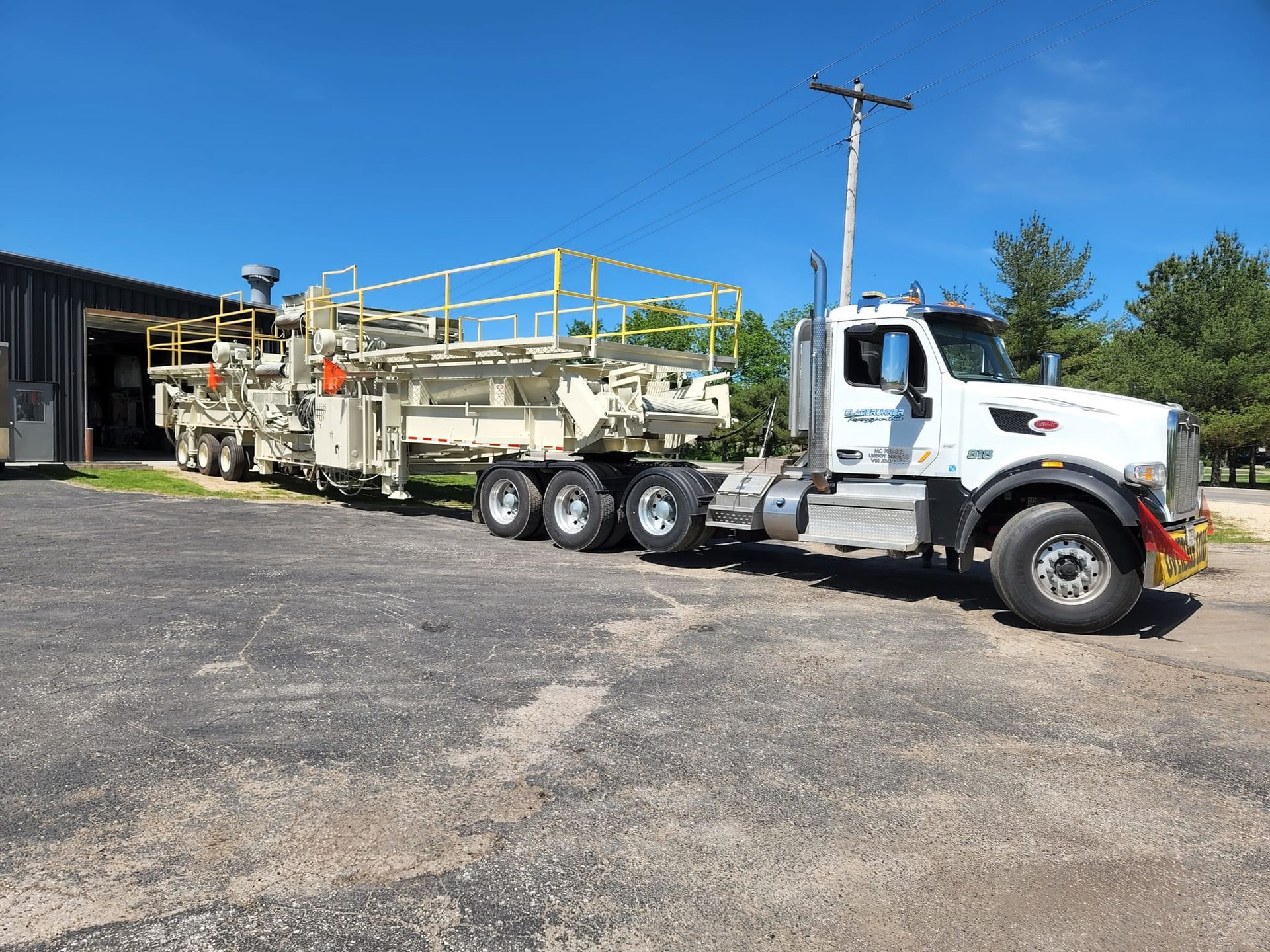 A white semi-truck pulls a large, beige industrial construction machine parked on an asphalt lot under a blue sky.