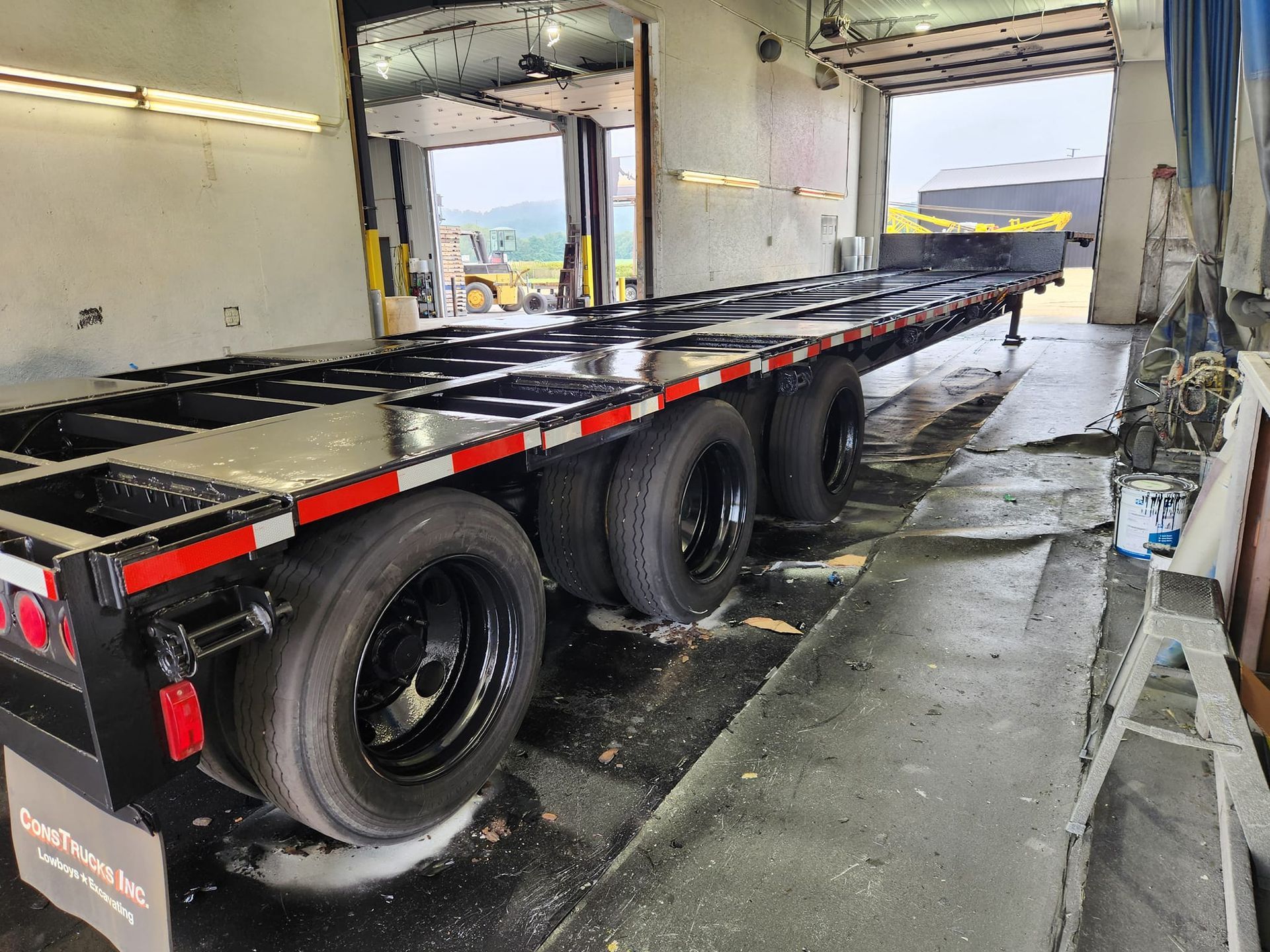 A long, black flatbed semi-trailer parked inside a maintenance bay with bright red reflective tape along its side.