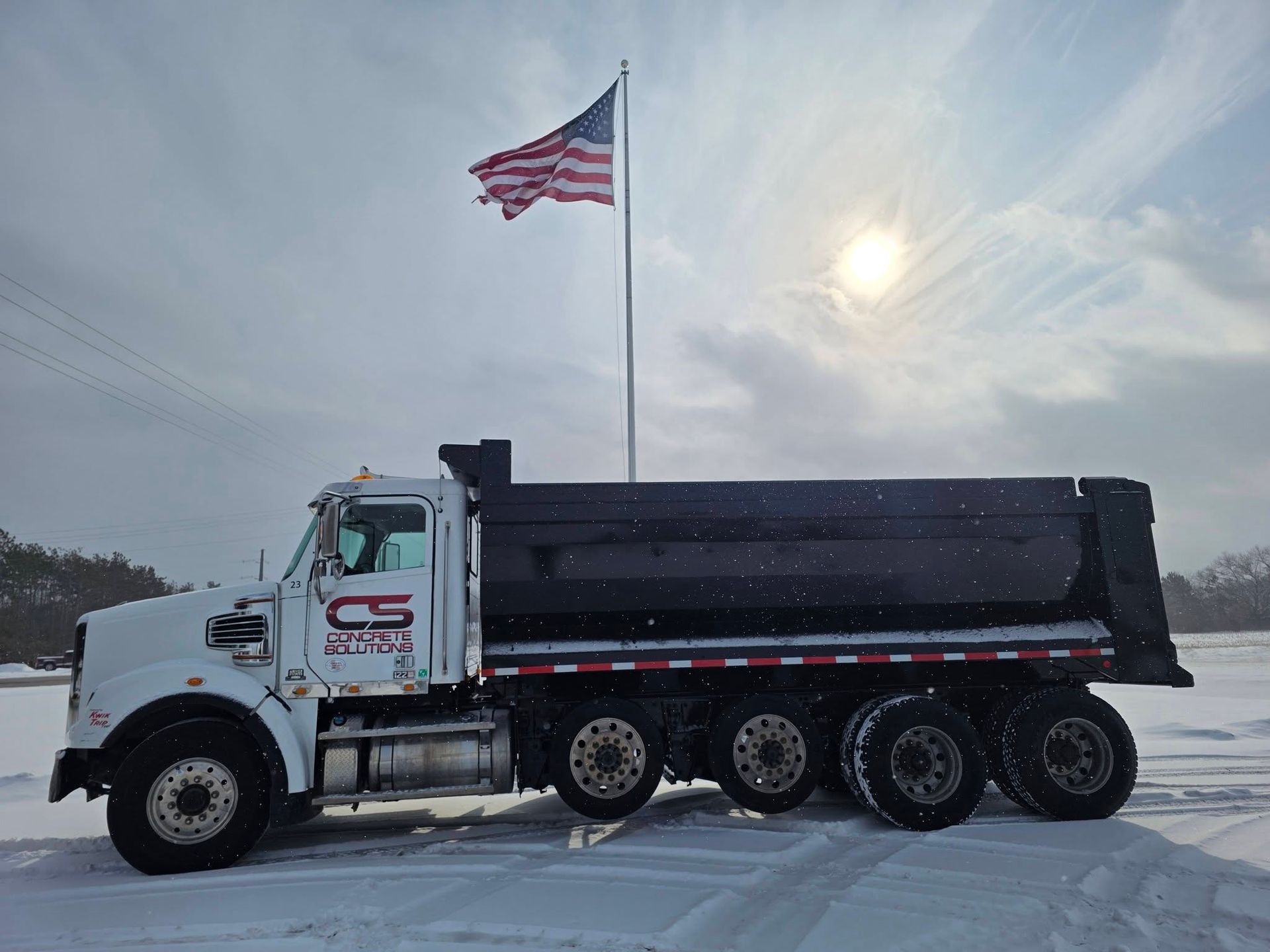 A white dump truck with a black cargo bed parked in a snowy field beneath a waving American flag.