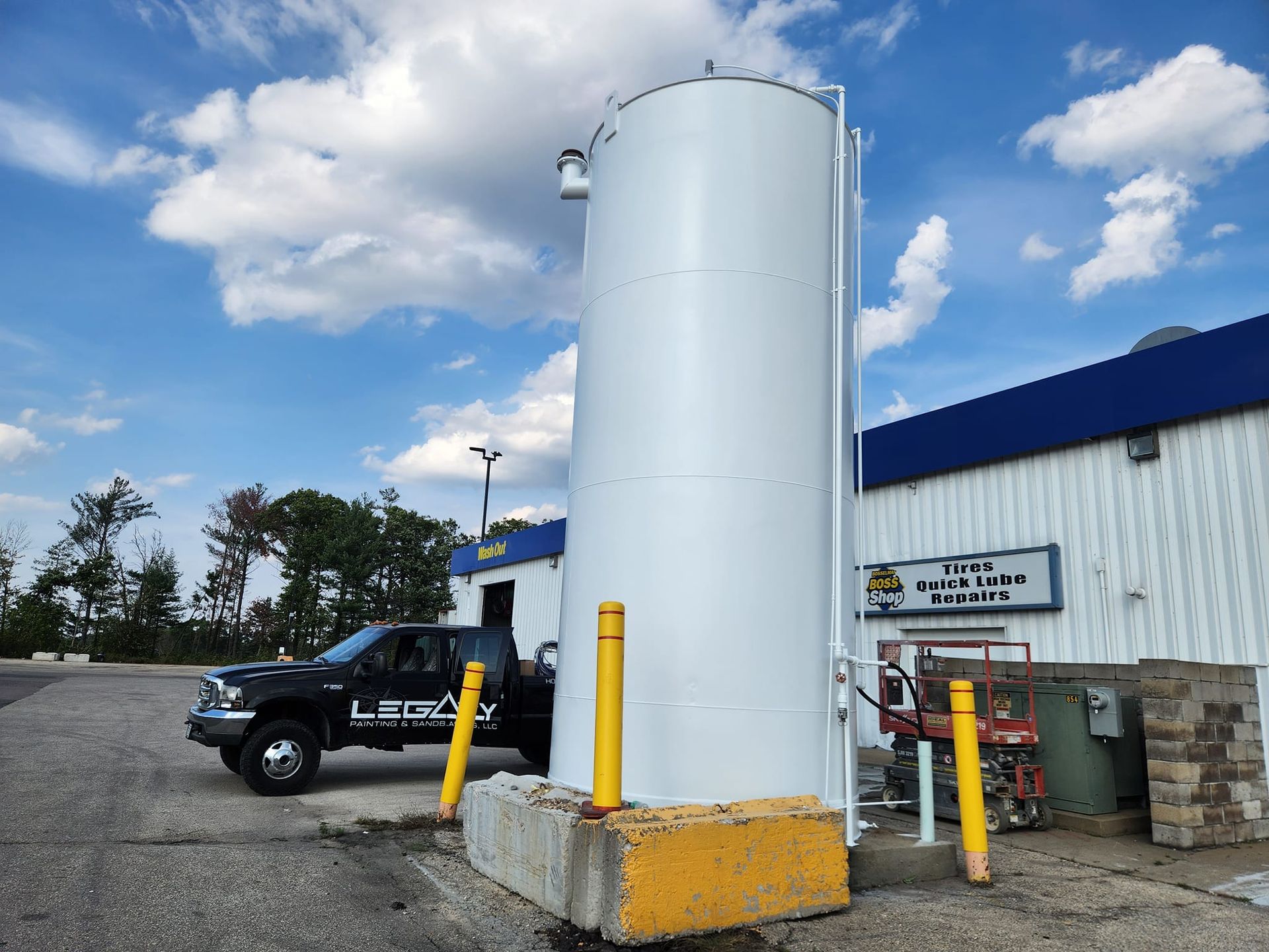 A tall, white vertical storage tank sits on a concrete base next to a black pickup truck and a building under a blue sky.