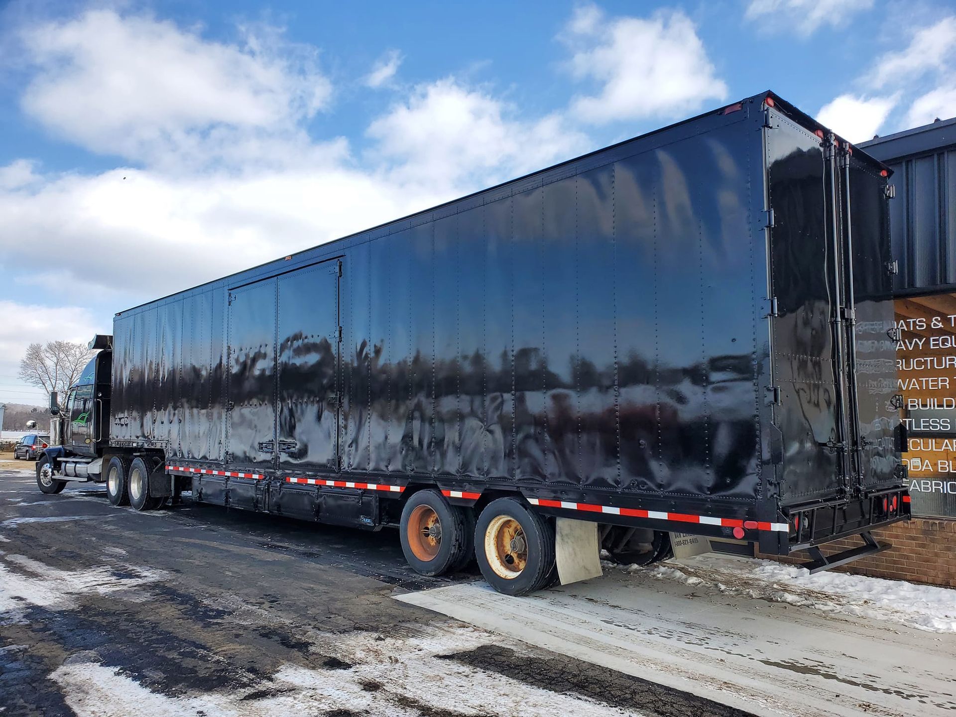 A long, black semi-trailer truck parked on a snowy road under a blue, cloudy sky.