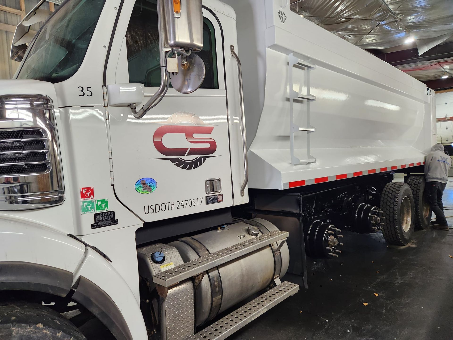 A white dump truck with a "CS" logo parked in an indoor garage, with a person standing near the rear tires.