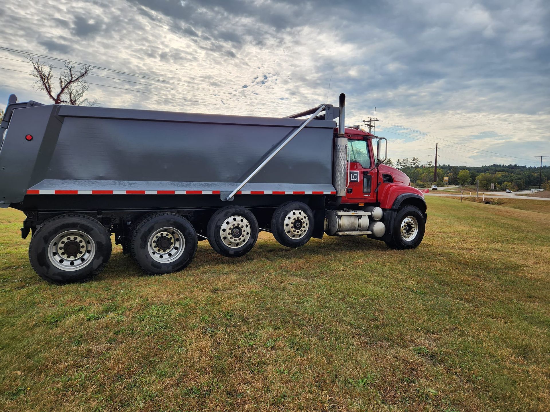 A red dump truck with a large, grey cargo bed parked on a grassy field under a cloudy sky.