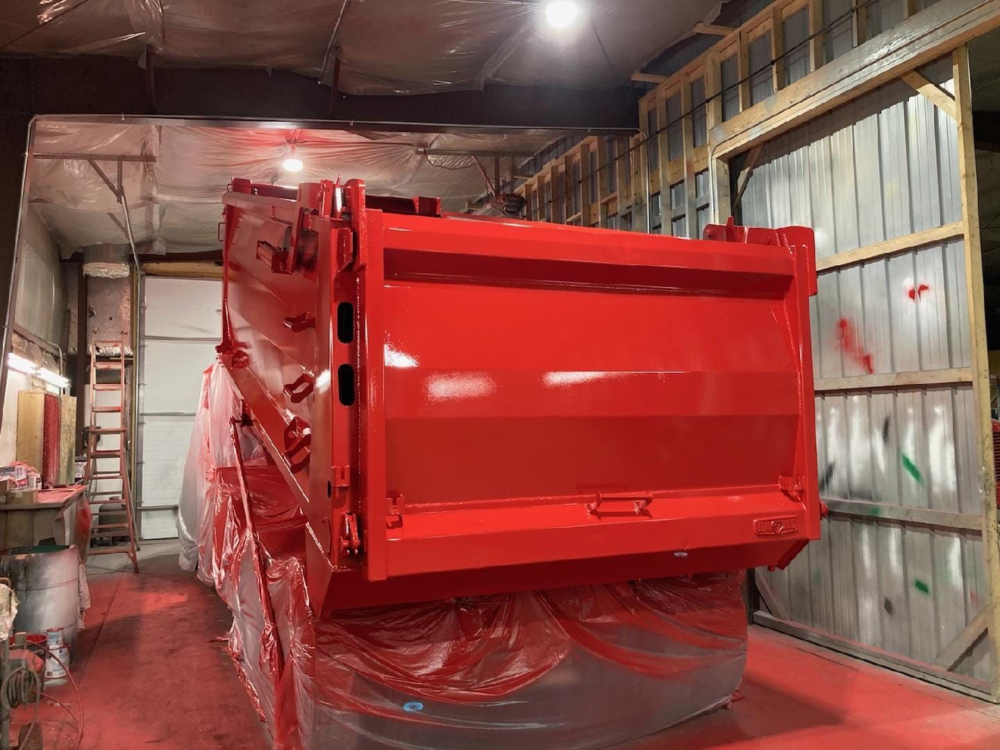 A freshly painted, bright red dump truck bed inside an industrial spray booth, partially covered in protective plastic.