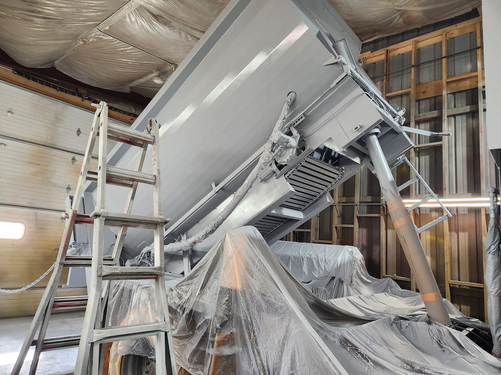 A large, grey-painted truck dump bed raised in a workshop, with a tall stepladder in the foreground.