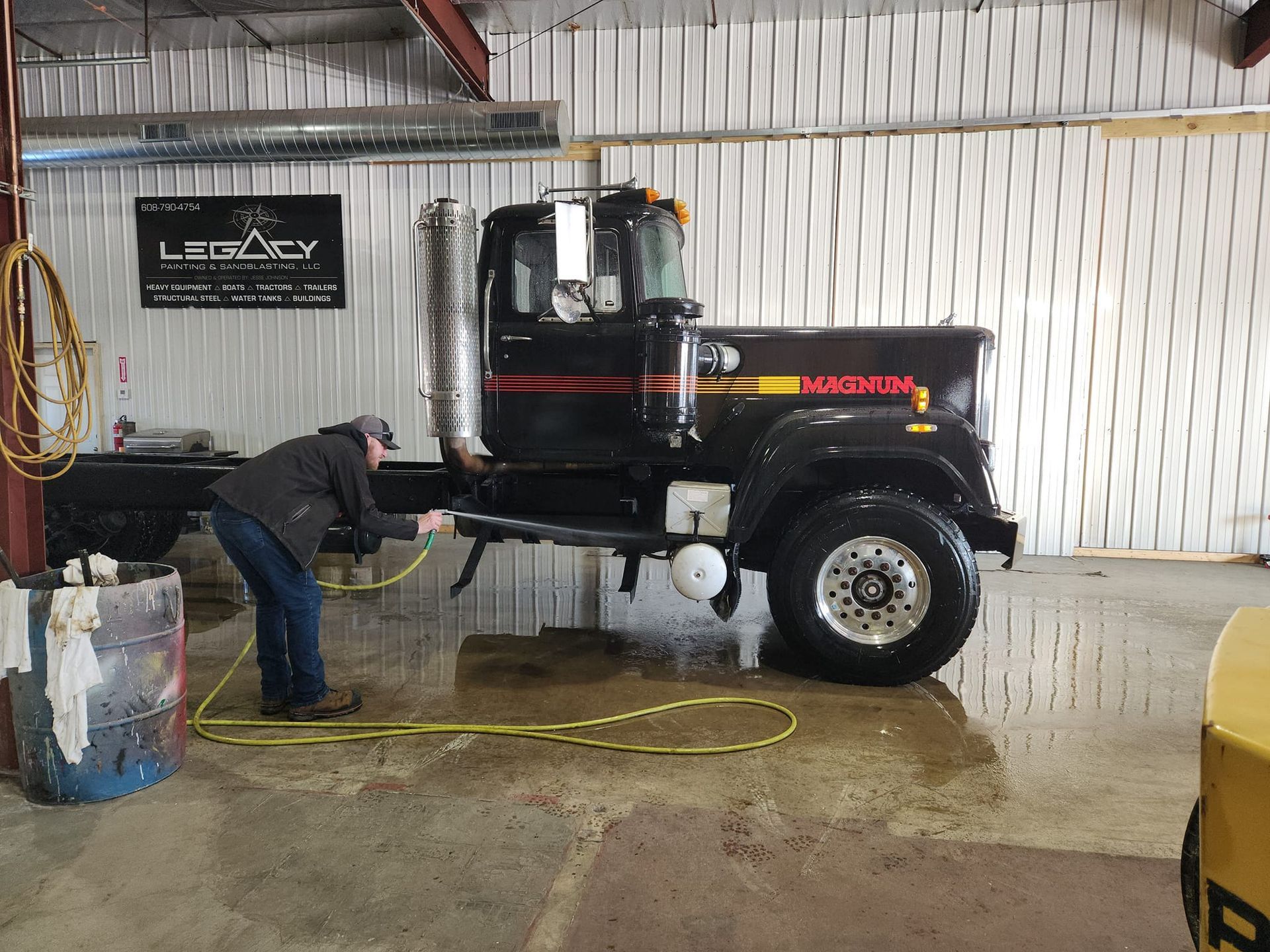 A worker sprays down a black semi-truck with a yellow hose inside a warehouse with a Legacy branding sign on the wall.