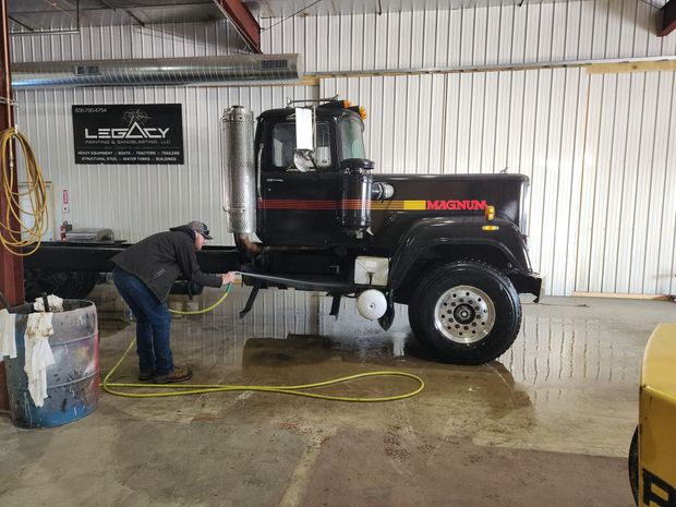 A worker sprays down a black semi-truck with a yellow hose inside a warehouse with a Legacy branding sign on the wall.