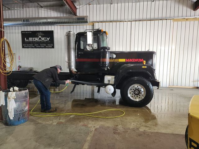 A worker sprays down a black semi-truck with a yellow hose inside a warehouse with a Legacy branding sign on the wall.