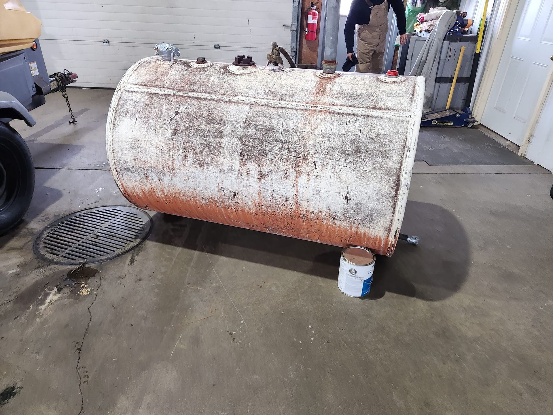A weathered, cylindrical white metal tank with rust sits on the floor of a workshop, propped up by a paint can.