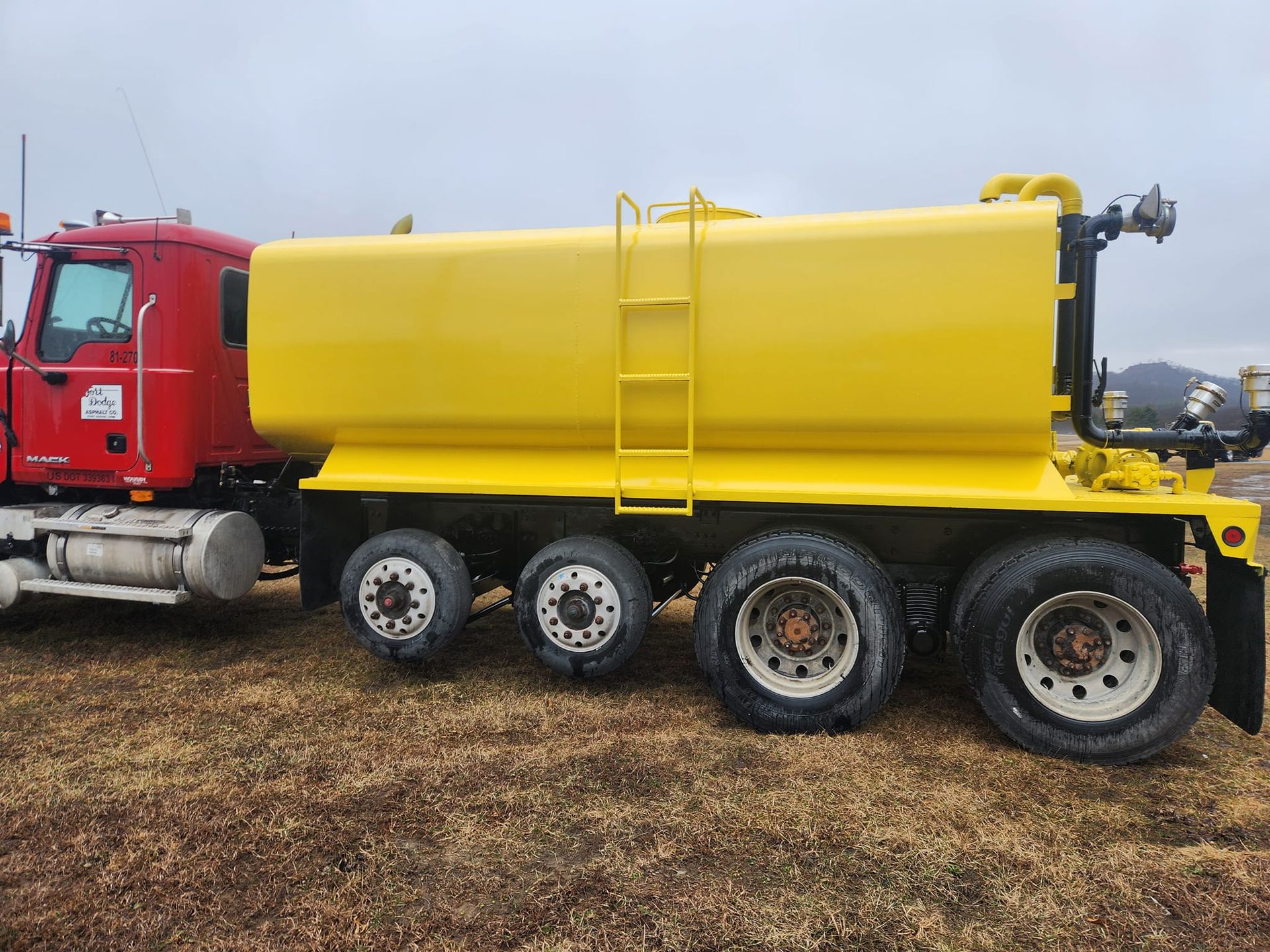 A red truck cab attached to a large yellow water tank trailer sitting on a grassy field under a cloudy sky.