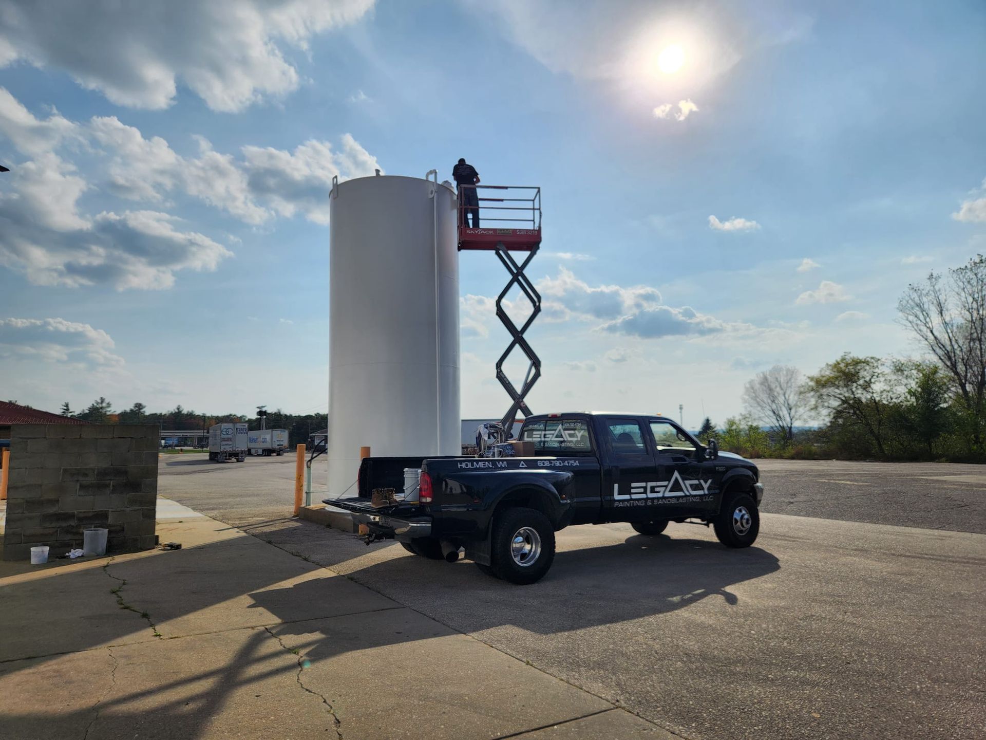 A worker on a scissor lift next to a tall white cylindrical tank, with a black pickup truck parked in an outdoor lot.