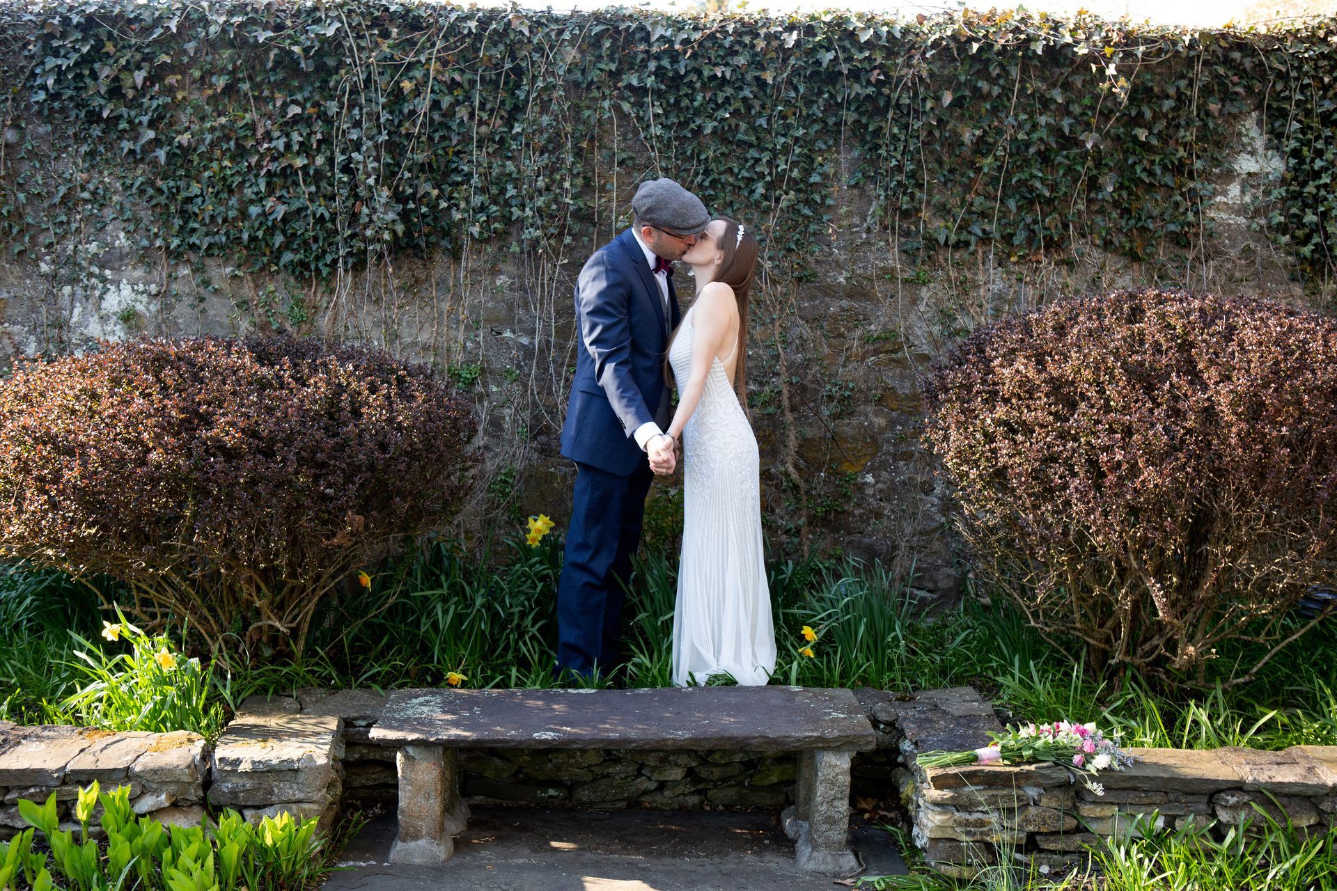 A bride and groom are kissing on a bench in a garden.