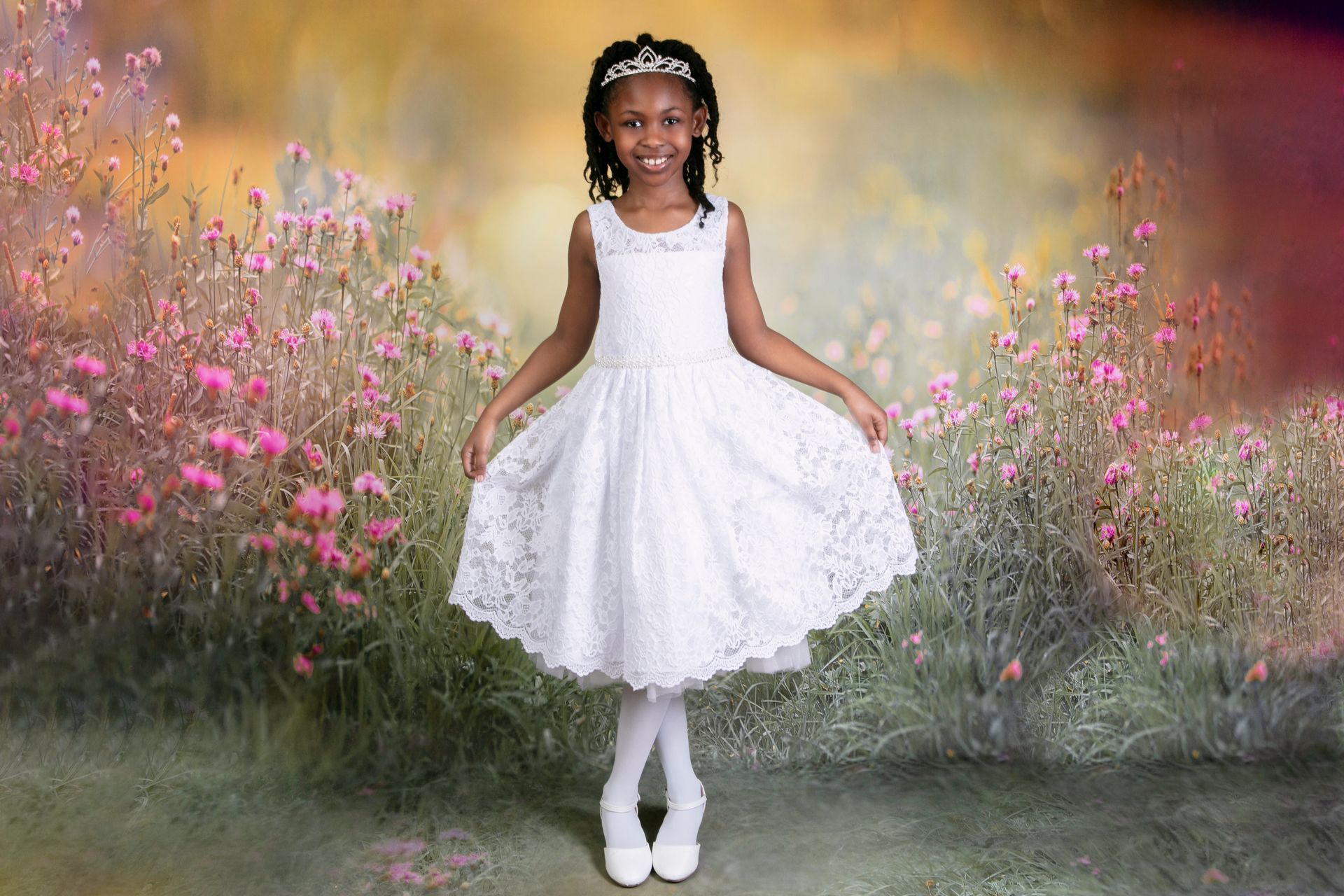 A little girl in a white dress and tiara is standing in a field of pink flowers.