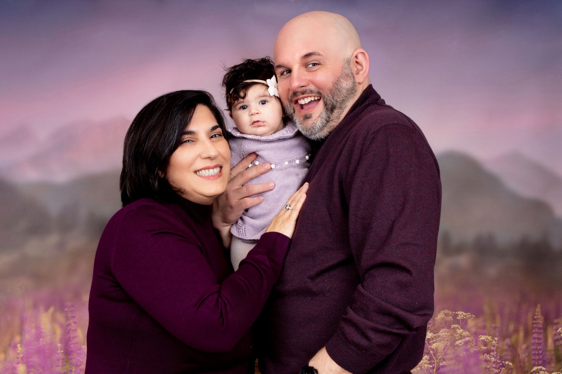 A man and woman are holding a baby in front of a purple background.