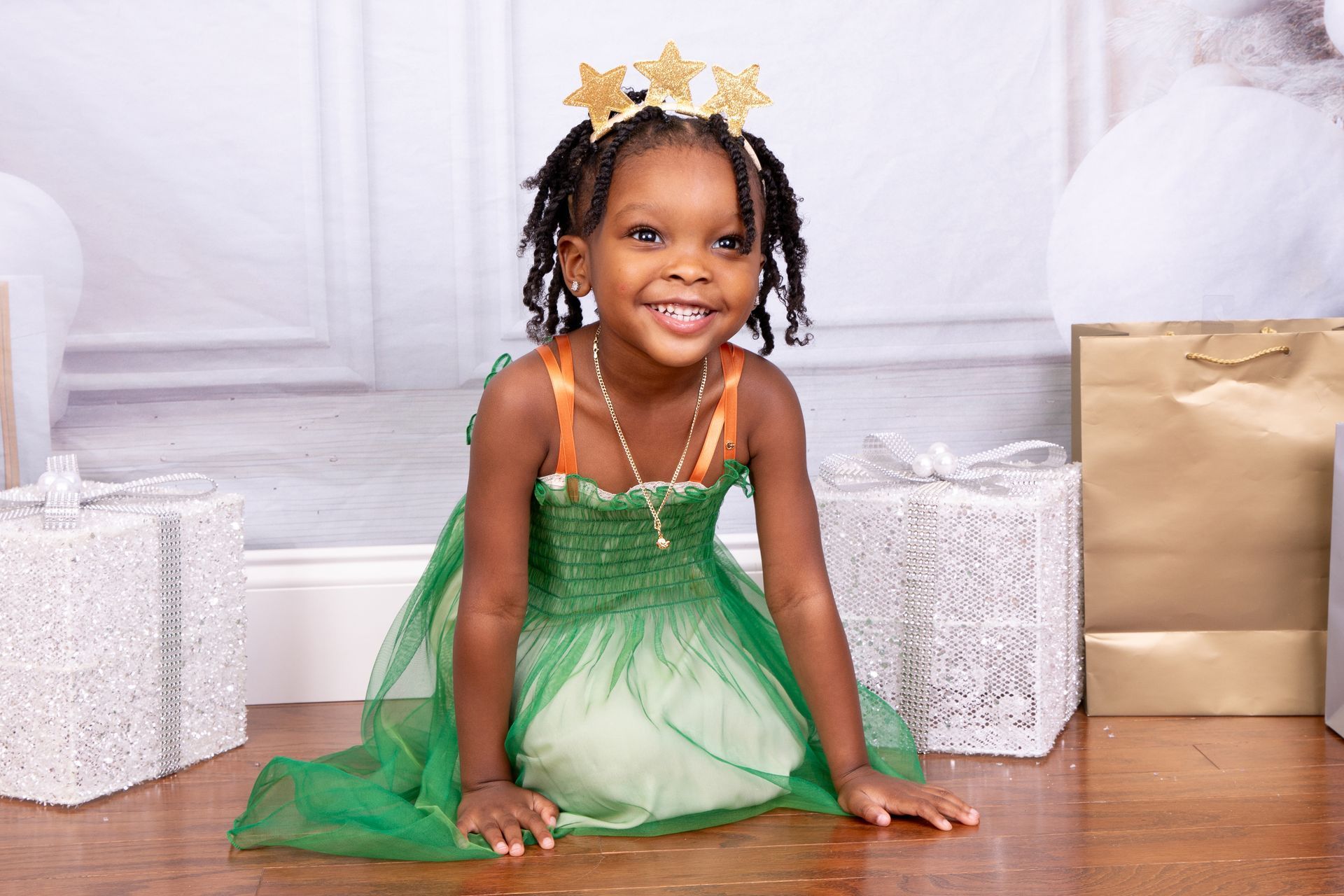 Smiling child in a green dress and gold crown, posed on a wood floor near wrapped gifts.