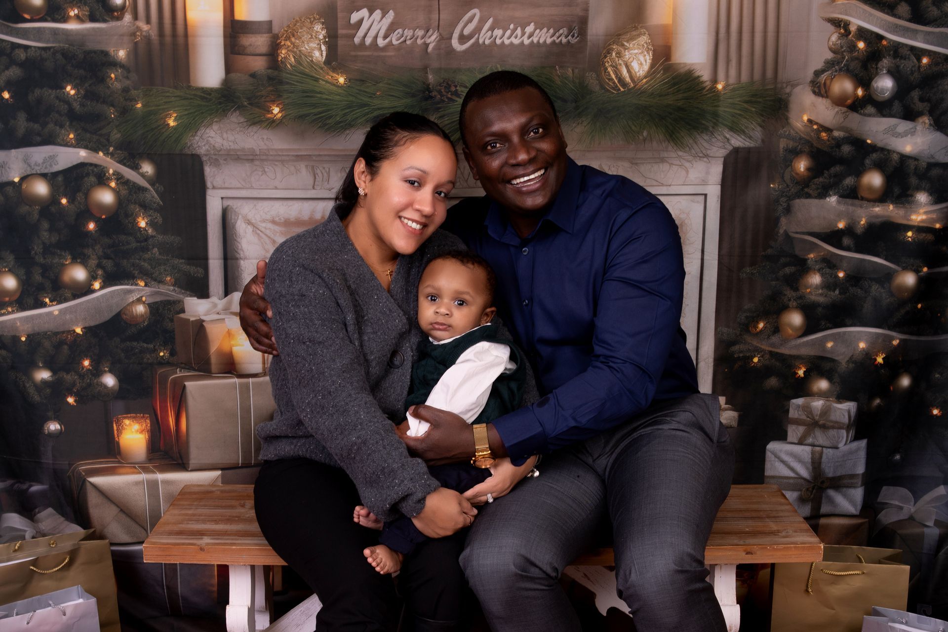 Family of three smiling, seated in front of a Christmas backdrop. Woman, man, and child posing together.