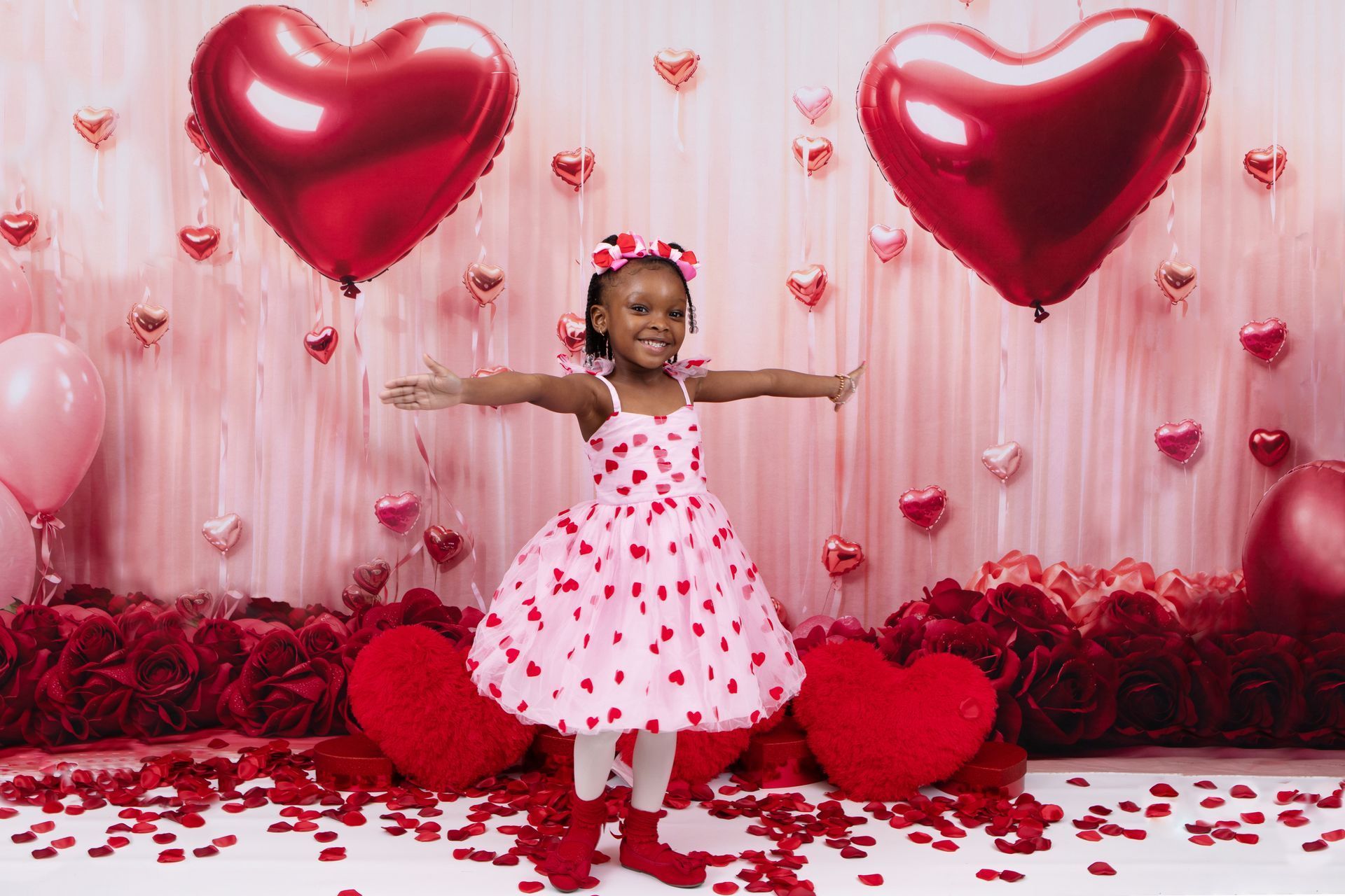 Girl in heart-print dress, arms outstretched, smiling among heart decorations and balloons.