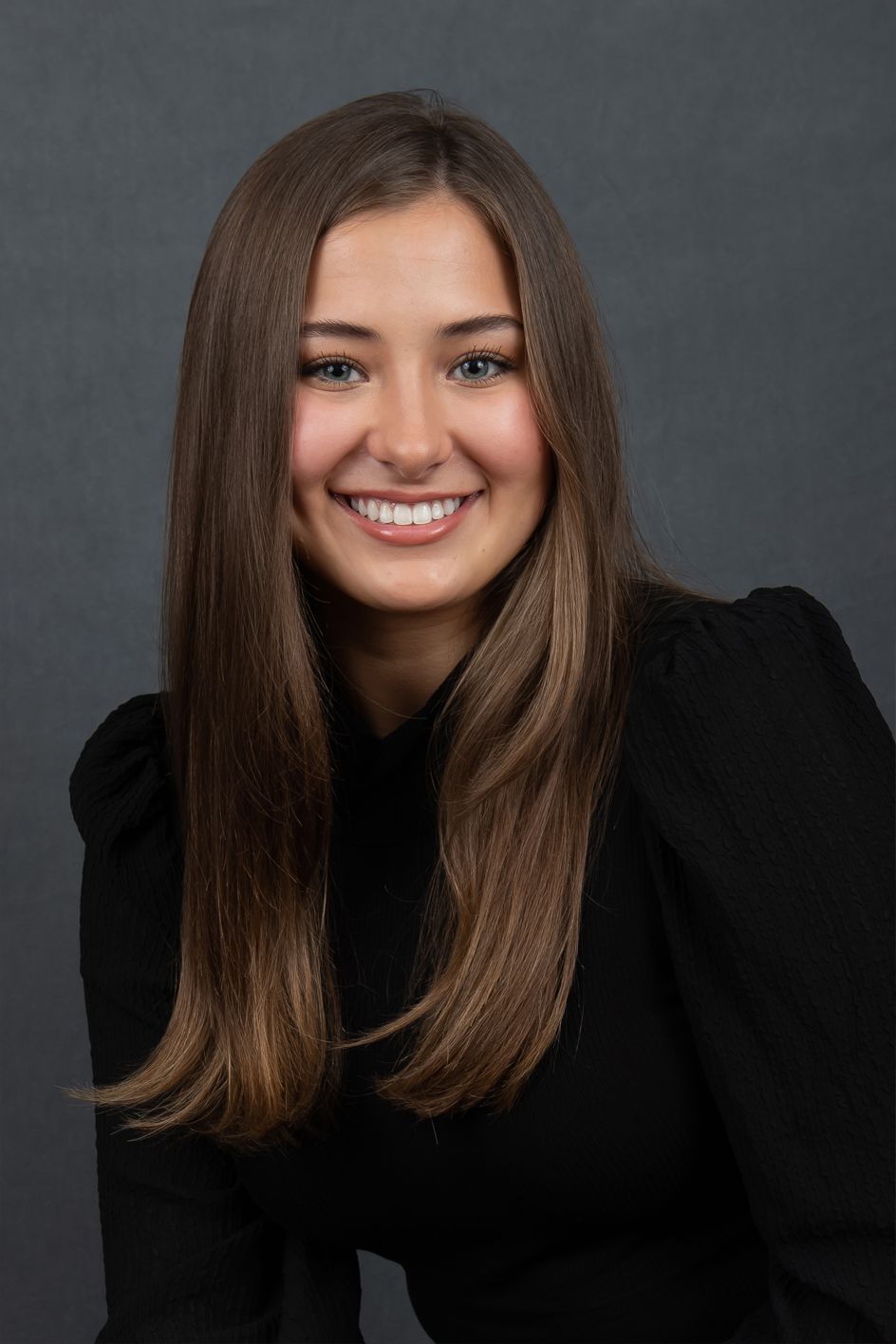 A woman with long brown hair is wearing a black shirt and smiling for the camera.