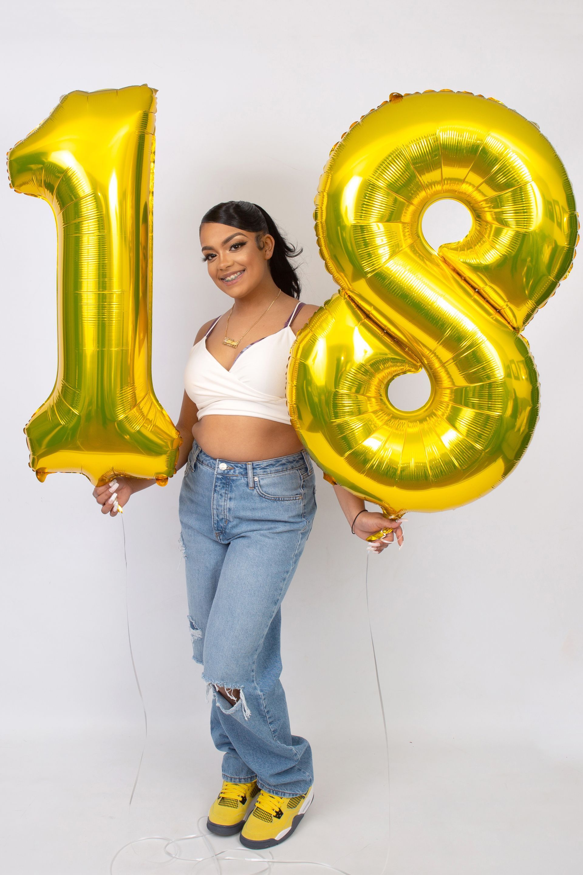 A woman is holding two gold balloons in the shape of the number 18.