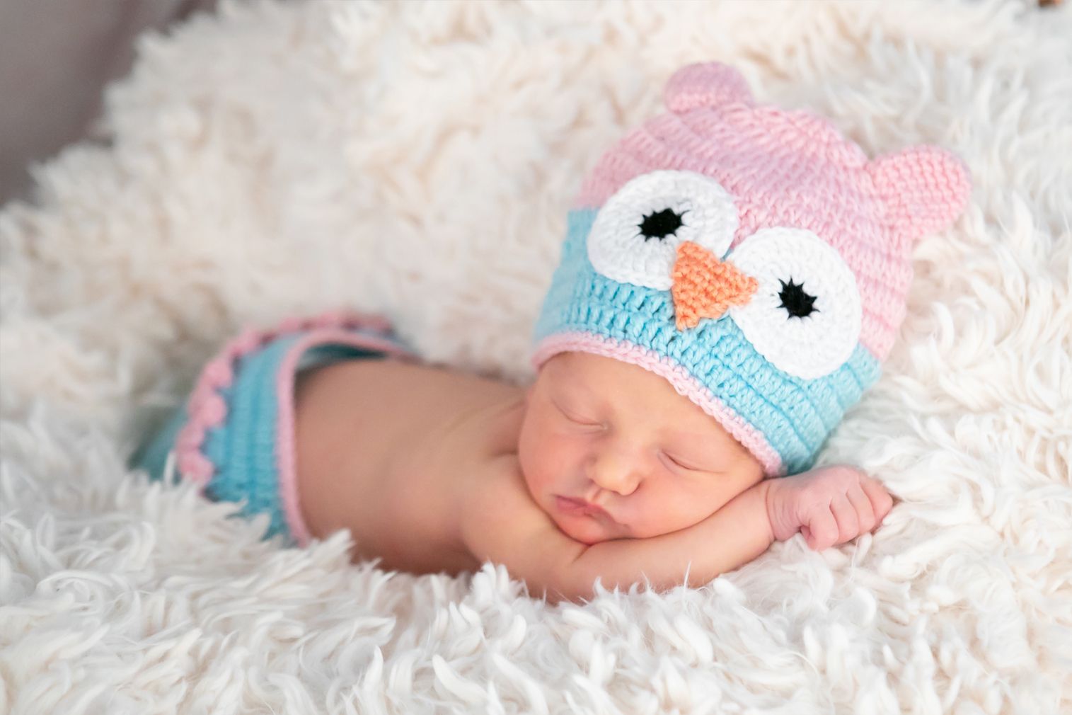 A newborn baby wearing a crocheted owl hat and pants is sleeping on a white blanket.