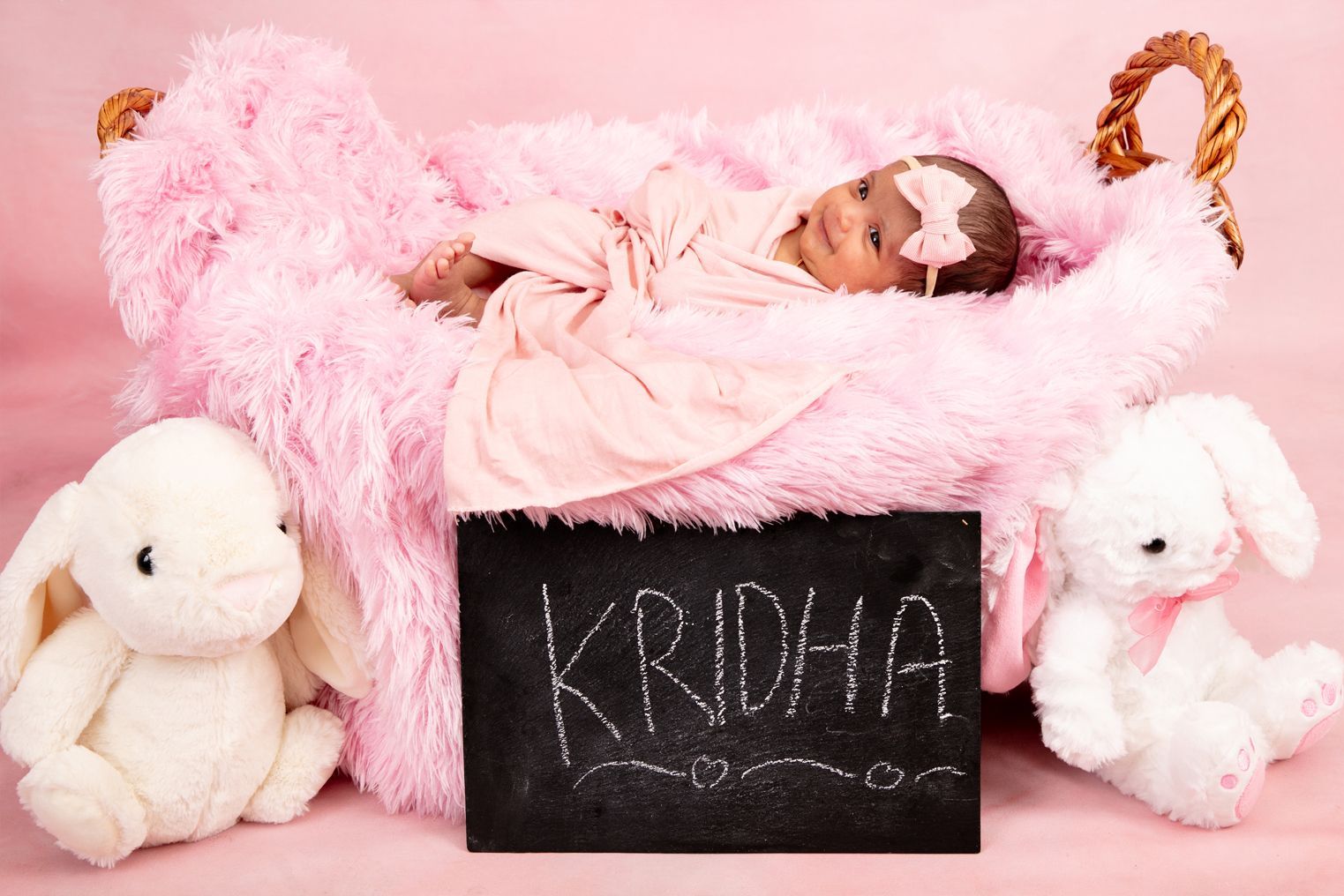 A baby is laying in a pink basket next to stuffed animals.