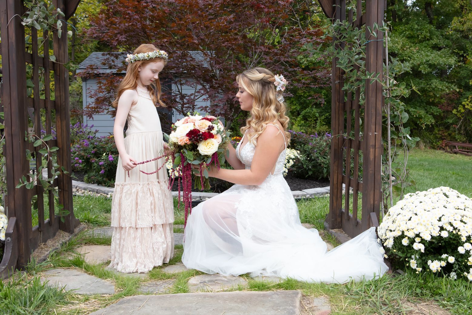 A bride is kneeling down next to a flower girl holding a bouquet of flowers.