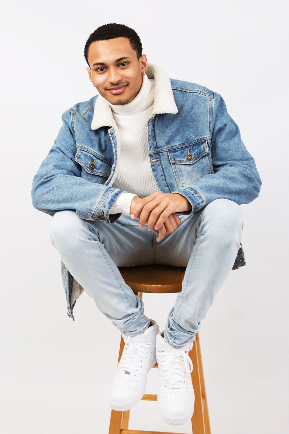 A man in a denim jacket and white turtleneck is sitting on a wooden stool.