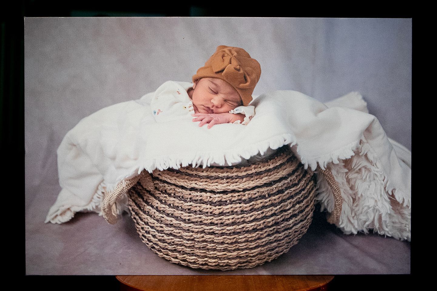Newborn baby sleeping peacefully in a woven basket, covered by a fringed white blanket and wearing a tan knit cap, on a neutral backdrop.