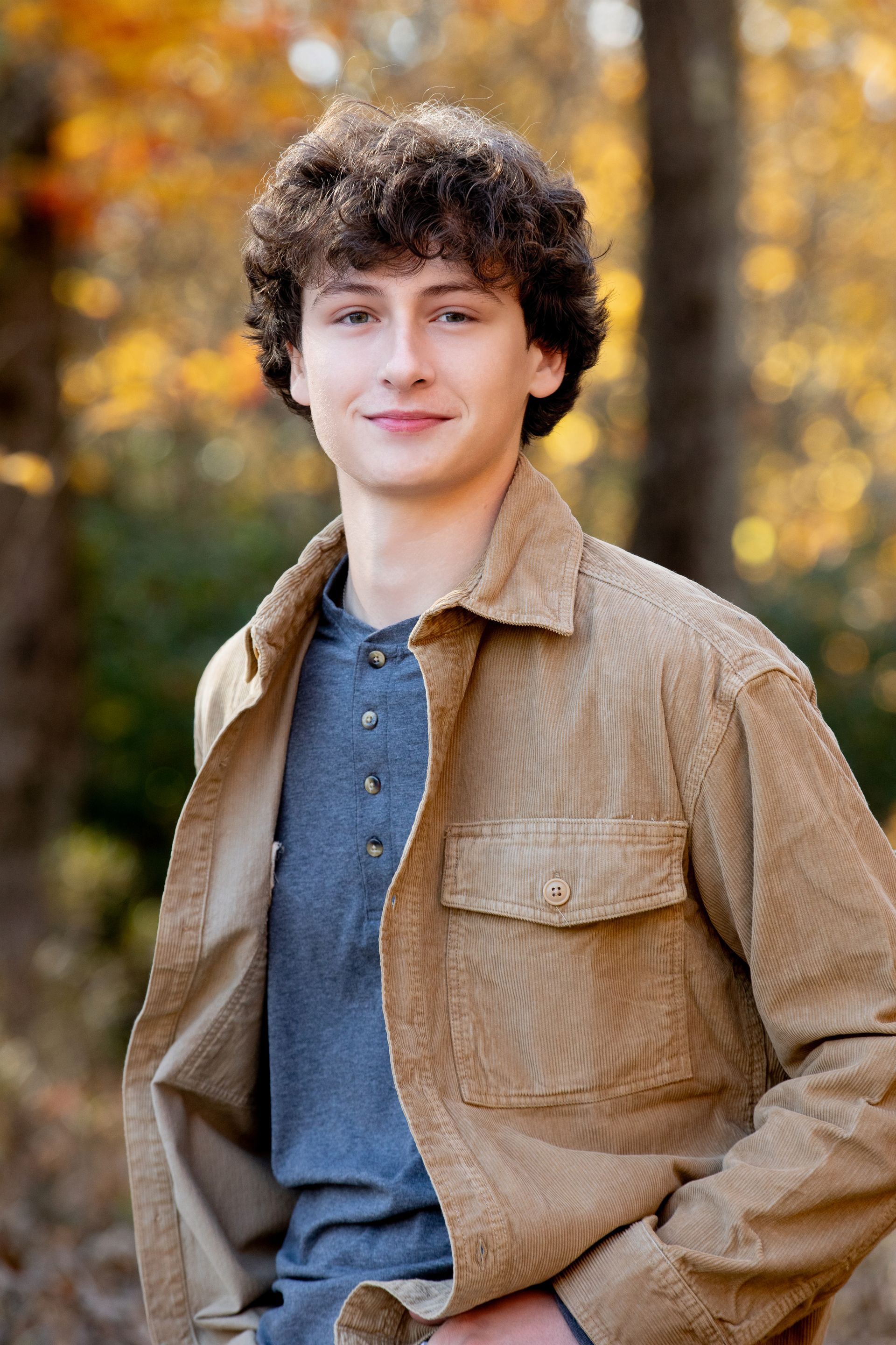 A young man in a brown jacket is standing in the woods.