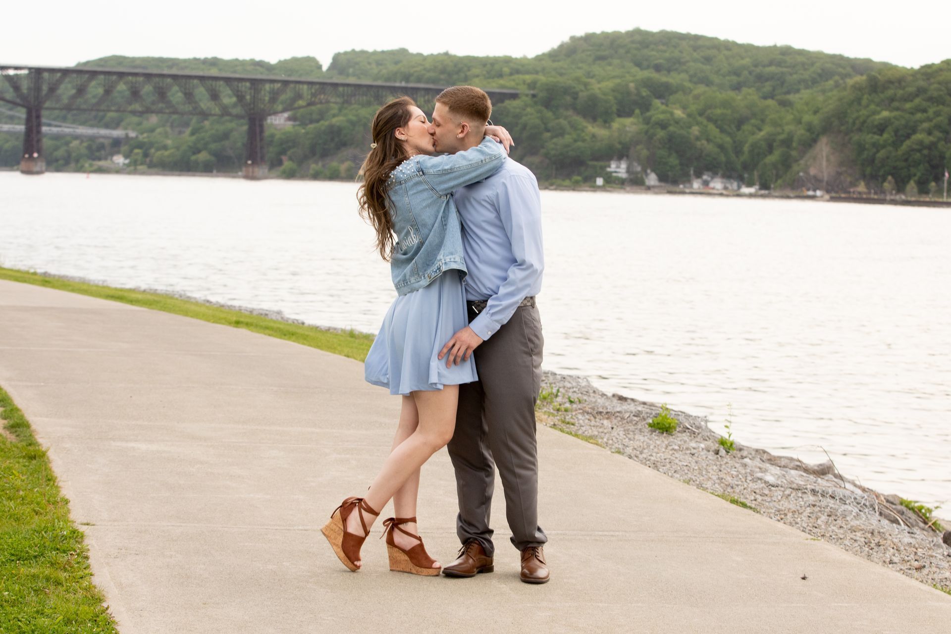 A man and a woman are kissing on a sidewalk next to a body of water.