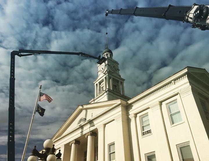 Cranes lifting object atop a white building with clock tower, American flag in foreground, cloudy sky.