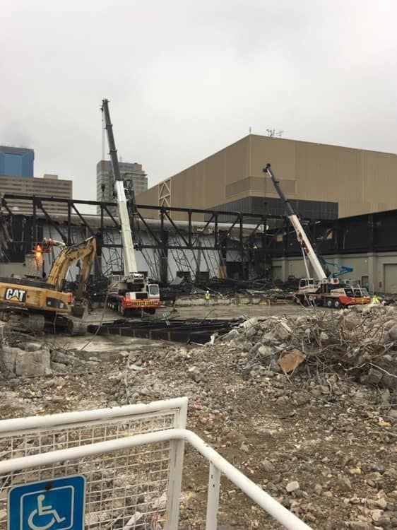 Construction site with cranes demolishing a black steel structure next to a large tan building.