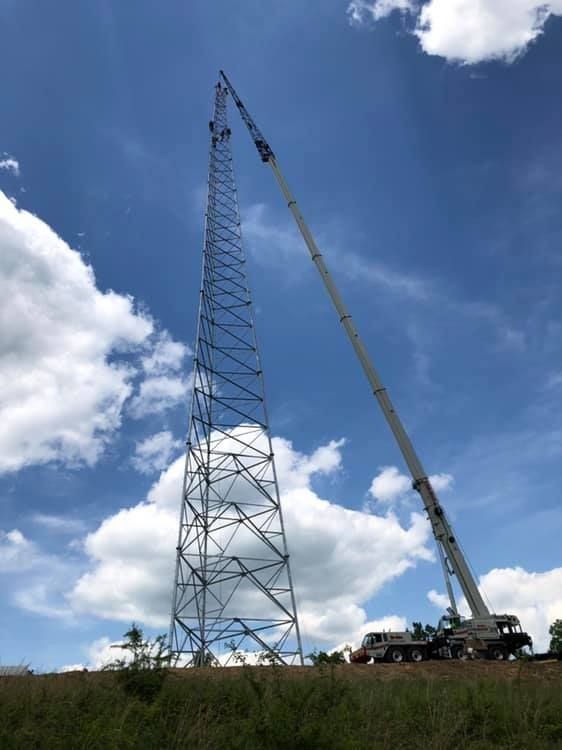 Crane assembling a tall, steel telecommunications tower on a grassy hill under a partly cloudy sky.